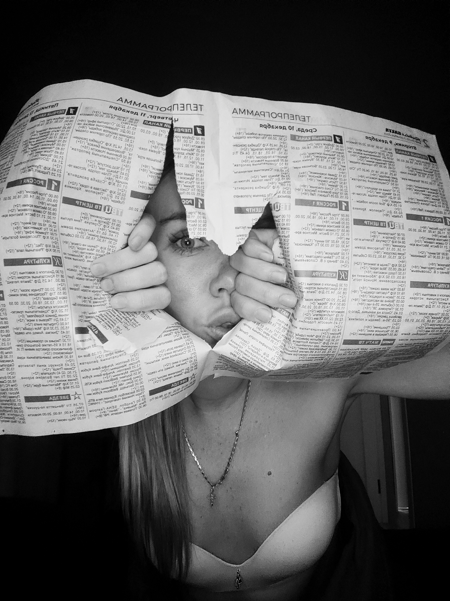 This black and white photograph captures a young woman peeking through a jagged, torn opening in a newspaper. Her face, primarily her wide, alert eyes, nose, and mouth, is centrally framed by the ripped paper. Her hands are visible, gripping the torn edges of the newspaper firmly. She has long, light-colored hair, part of which is visible over her shoulders. Around her neck, she wears a delicate chain necklace with a small cross pendant.
The newspaper is covered in Cyrillic text and numbers, indicative of broadcast schedules or articles. Numerous age ratings such as "(+0)", "(+6)", "(+12)", and "(+18)" are present. Prominent visible words include "АММАРГОТЕПЕП" (likely mirrored or reversed, possibly related to TV programs), "РОССИЯ" (Russia), "СВЯЗЬ" (Communication), and "АДРЕСА" (Addresses). Some sections of the text appear right-side up, while others are inverted or mirrored, suggesting how the paper is held.
Below the newspaper, the woman's shoulder and upper chest are visible, indicating she is wearing a bra or similar undergarment. The background is dark and indistinct, hinting at an indoor setting, with a faint reflection on the lower right suggesting a door frame. The image conveys a sense of hidden observation or playful curiosity. The Cyrillic text is consistent with the provided location context of Votkinsk, Russia.