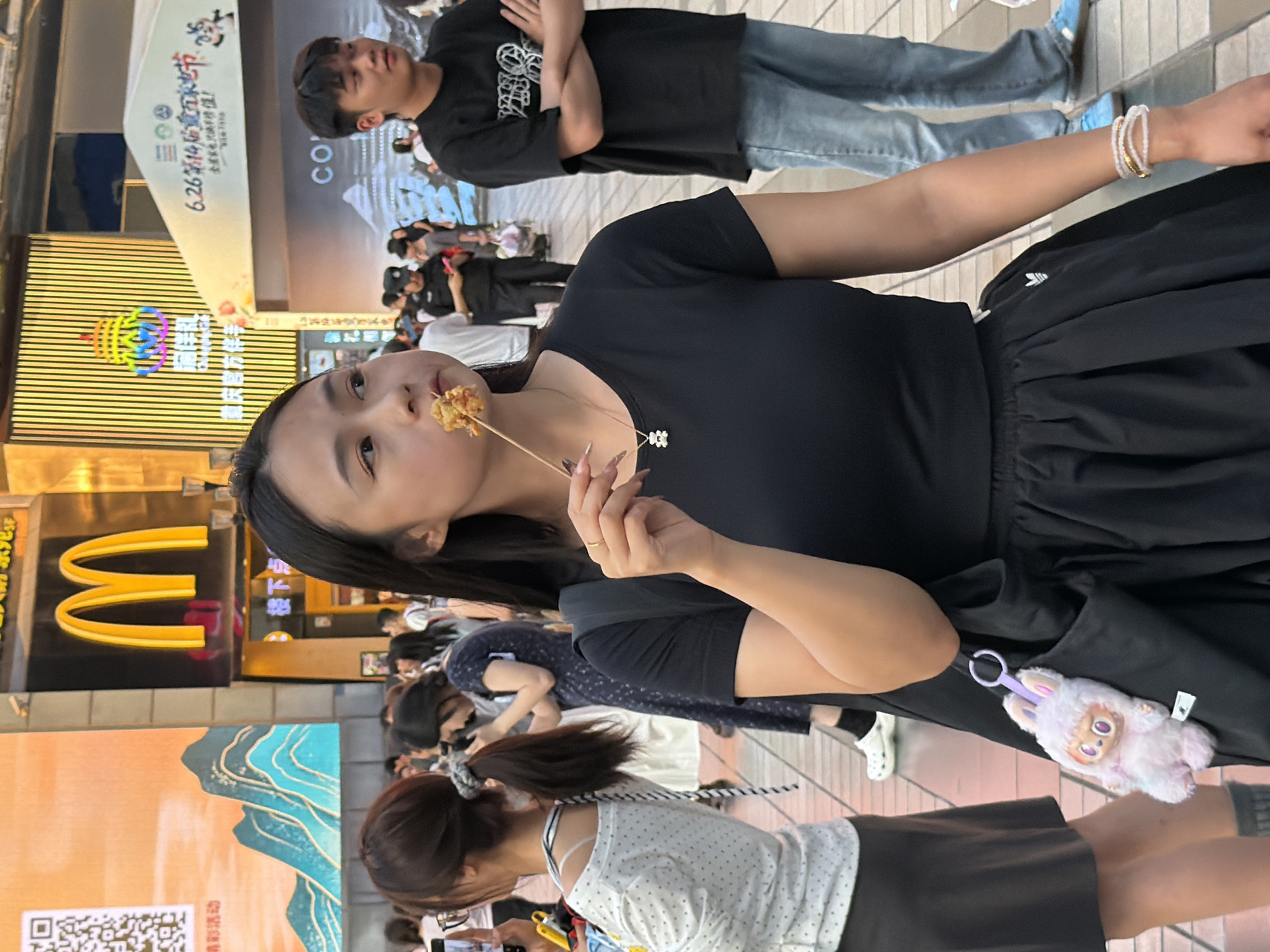 In the Yuzhong District of China, a young woman with dark hair is featured in the foreground, holding a skewer with a piece of food to her mouth. She wears a black crop top, a black skirt, and a necklace with a white pendant. A black bag hangs from her shoulder, adorned with a fluffy pink bunny keychain. To her right, a young man in a black t-shirt and jeans stands with his arms crossed. In the background, a McDonald's sign is visible, along with other people walking in a public area. The environment appears to be a bustling street or plaza.