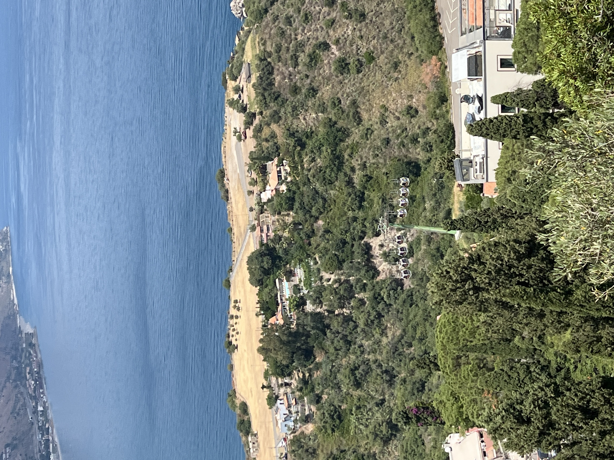 This is an aerial, high-angle, daylight, eye-level shot of Taormina, Sicily, Italy. A body of water, likely the Ionian Sea, dominates the upper half of the image, with a coastline visible in the upper left. The middle ground features a steep, tree-covered hillside with several buildings and a cable car system ascending the slope. The cable car, a common mode of transport in Taormina, has multiple cabins visible. The foreground shows lush green trees and foliage, with more buildings partially visible. The scene captures the characteristic Mediterranean landscape and architecture of Taormina, a popular tourist destination known for its dramatic coastal views and historical sites.
