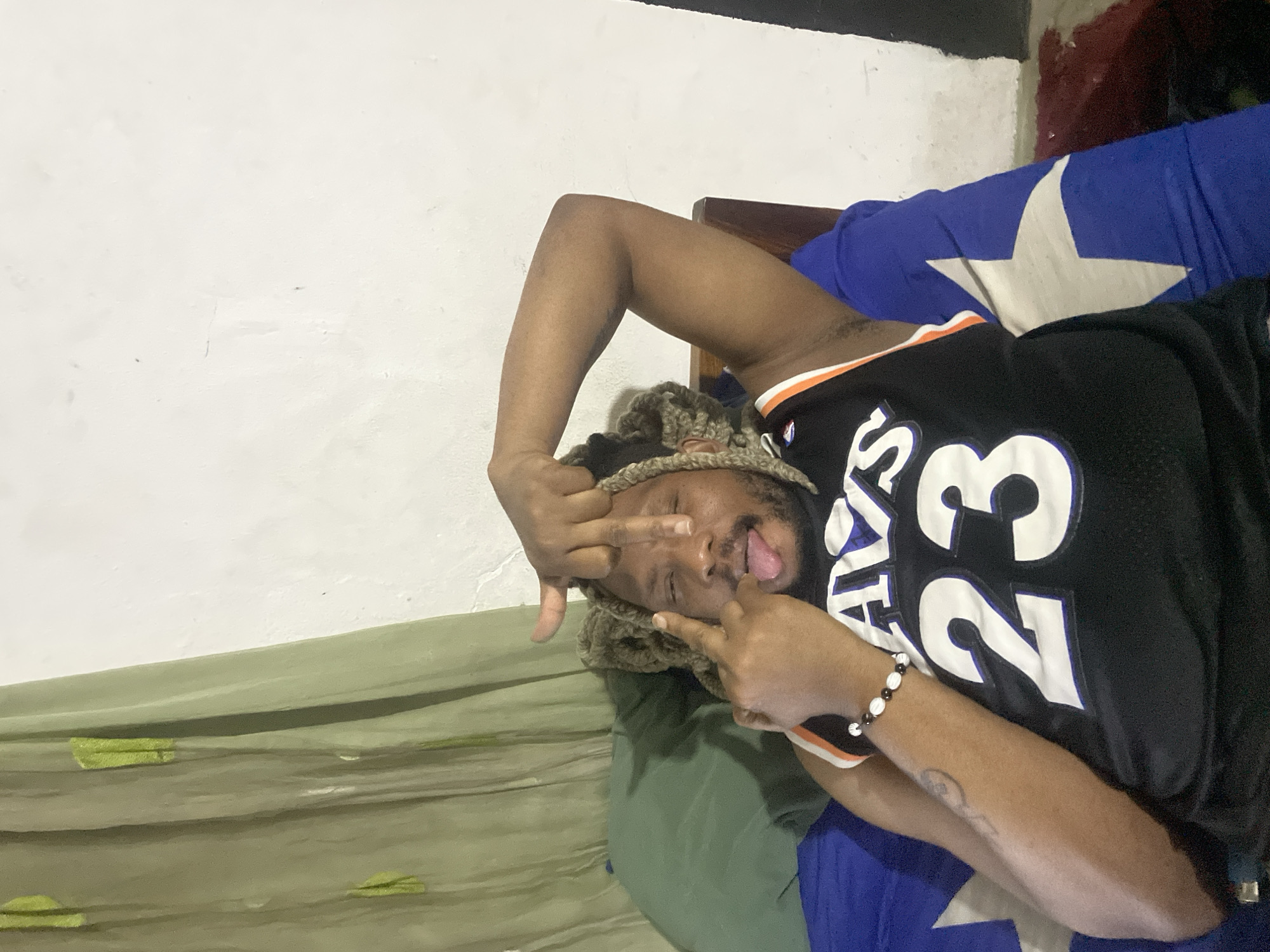 A Black man wearing a black jersey with the number "23" is lying on a bed with blue and green bedding. He is wearing a crocheted hat and has a bracelet on his right wrist. He is making a rude gesture with both hands while sticking out his tongue. The background shows a white wall and a dark area above the bed. The location is presumed to be in Jos, Nigeria.
