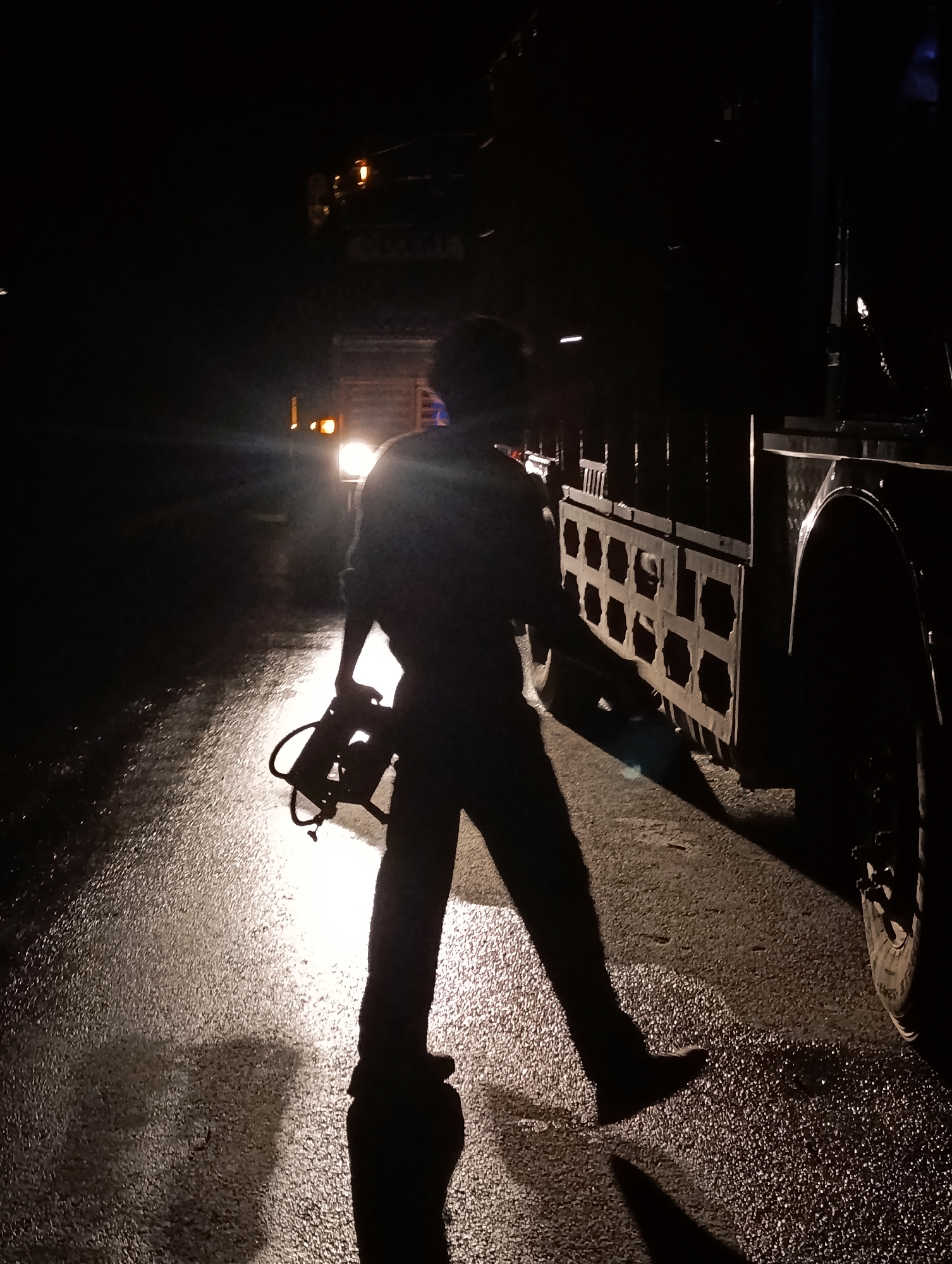 A silhouette of a person holding a portable power tool stands on a wet, reflective asphalt surface at night. The person is facing away from the camera and is lit from behind by the headlights of a vehicle. In the background, another vehicle, possibly a truck, has its headlights illuminated. The scene appears to be a road in Gemur, India.