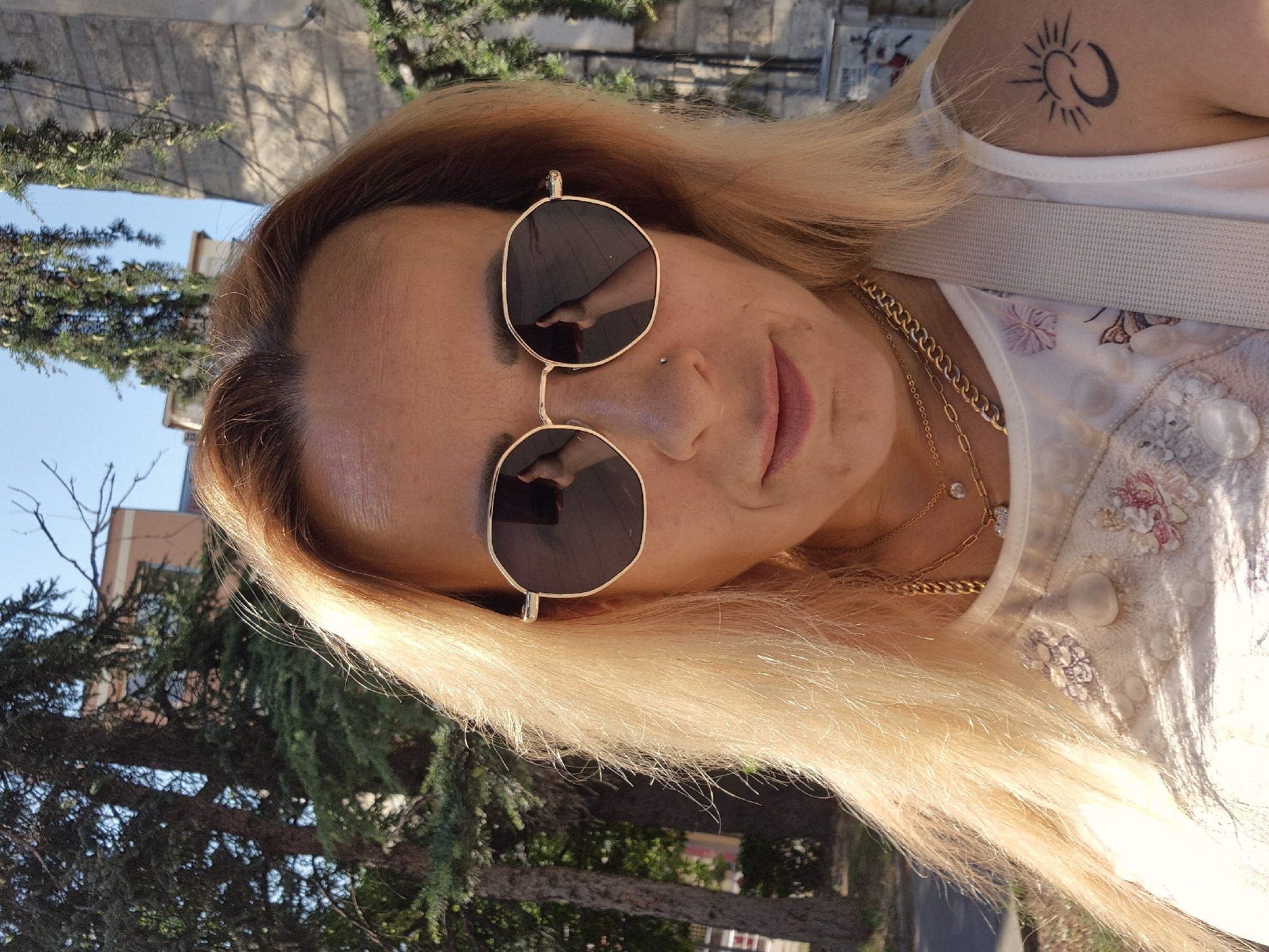 A woman with blonde hair and sunglasses is pictured outdoors, in what appears to be a city setting. She is wearing a white top with floral embroidery and multiple necklaces. A tattoo of a sun and crescent moon is visible on her upper left arm. The background features trees, a building, and a stone structure. The lighting suggests it is daytime.