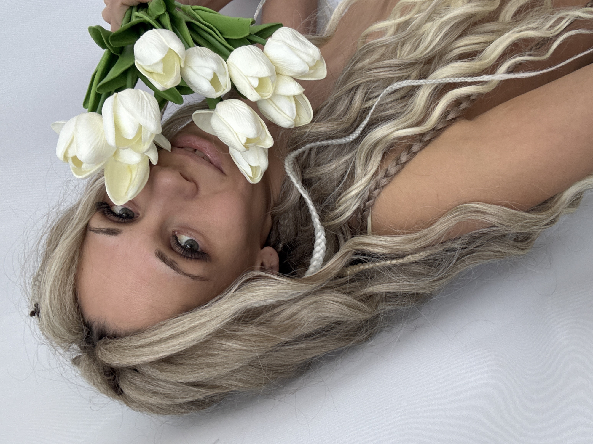 A woman with long blonde wavy hair is lying on a white surface, holding a bouquet of white tulips near her face. She is positioned at an angle, with her head tilted. Her facial features are visible, including her eyes, nose, and lips. She is wearing false eyelashes. Braided strands of white material are visible in her hair. The background is a plain white. It is difficult to ascertain if this image was taken in Rusanovo, Russia based on the provided information.
