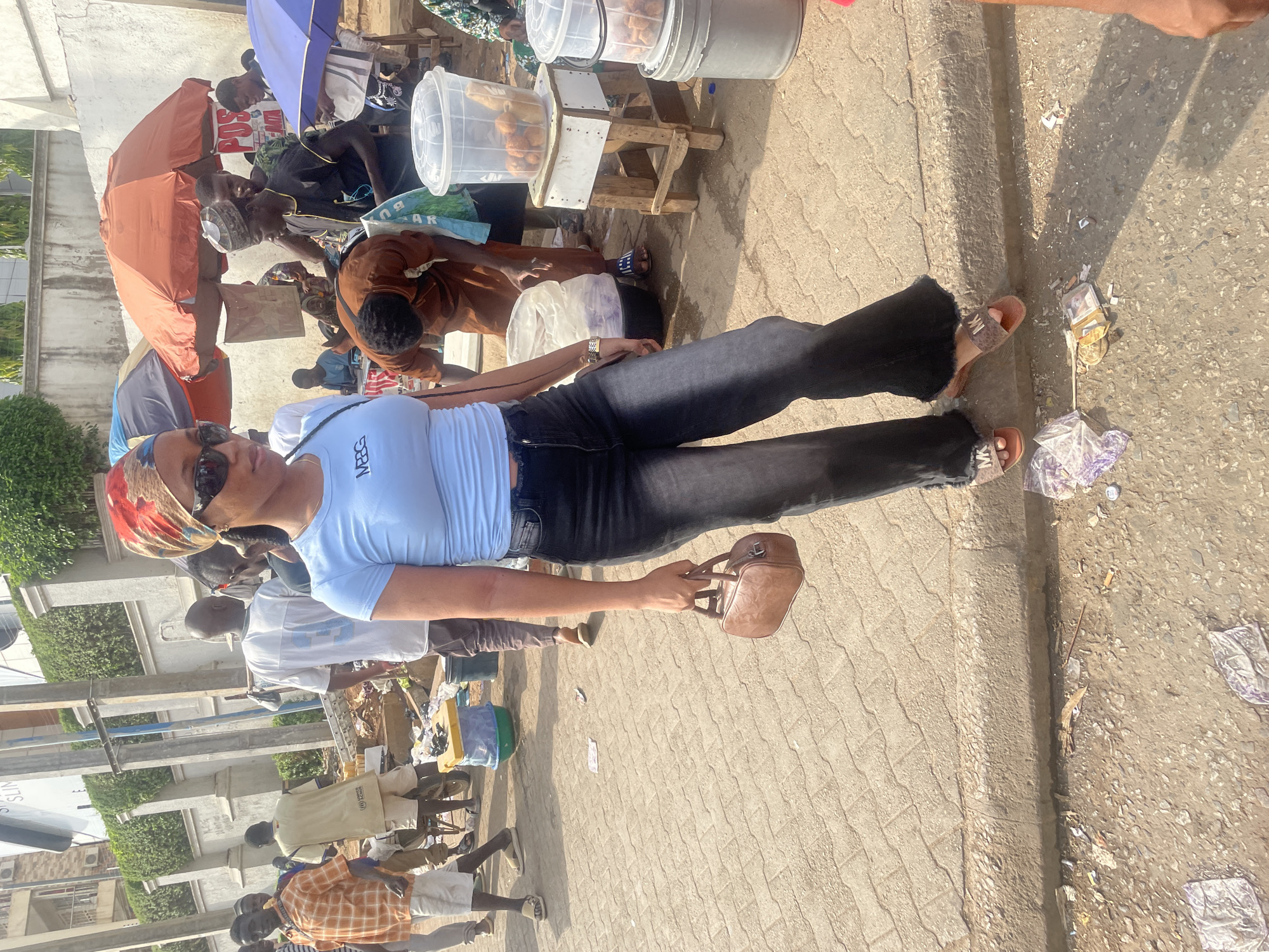 The image captures a bustling street scene, likely a market, in Abuja, Nigeria, under bright daylight. In the foreground, a woman stands upright, facing slightly to her left. She wears dark sunglasses, a colorful patterned headscarf (red, orange, brown, white), a light blue short-sleeved top with "MRG" printed on the chest, and dark blue or black jeans with frayed hems. Her feet are clad in brown flat sandals with "MK" visible on the strap. She holds a small, brown handbag in her right hand.
Around her, numerous other people are visible, many appearing to be vendors or customers. Several small stalls line the street, some shaded by large umbrellas, including a prominent orange one and a blue one further back. On wooden stands, clear plastic containers filled with what appear to be food items (possibly puff-puffs or similar fried snacks) are displayed for sale. One person in a brown shirt is bending over near a stall. The ground in the immediate foreground is paved with interlocking concrete blocks, while further to the right, it becomes a rougher, dirtier surface with visible debris, including wrappers and some scattered currency notes.
The bright sunlight casts strong shadows, indicating it is daytime, likely mid-morning or early afternoon. Visible text includes "MRG" on the main subject's shirt, "MK" on her sandals, "POS" and partially "DA...TA" on a sign under the orange umbrella, and "AR" on a blue bag near a stall. In the far left background, a sign partially shows "SUN." The scene conveys a lively, everyday atmosphere of commerce and activity.
