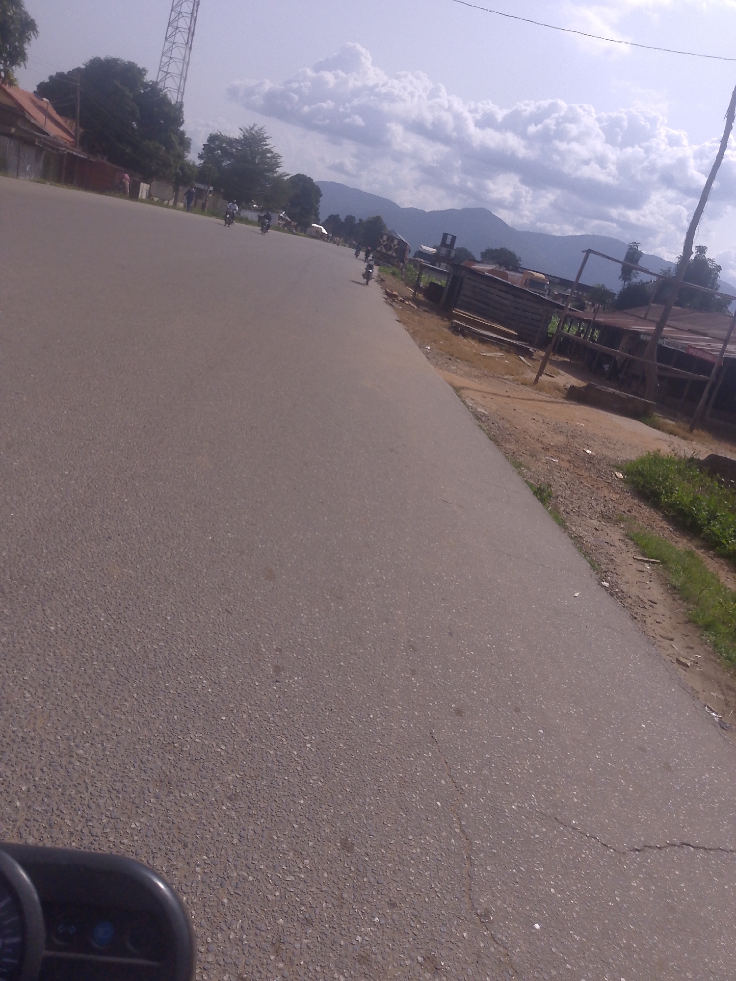 The image depicts a view from a moving vehicle, likely a motorcycle, on a paved road in Serti, Nigeria. The road is a dual carriageway with a slight incline. To the left of the road, a building with a red roof and white walls is visible, accompanied by trees and a tall radio tower. Several motorcycles are seen on the road, some with riders. On the right side of the road, there are wooden structures that appear to be shops or market stalls, with scattered debris on the ground. In the background, a range of mountains under a cloudy sky can be observed. The foreground shows a part of the vehicle's dashboard.
