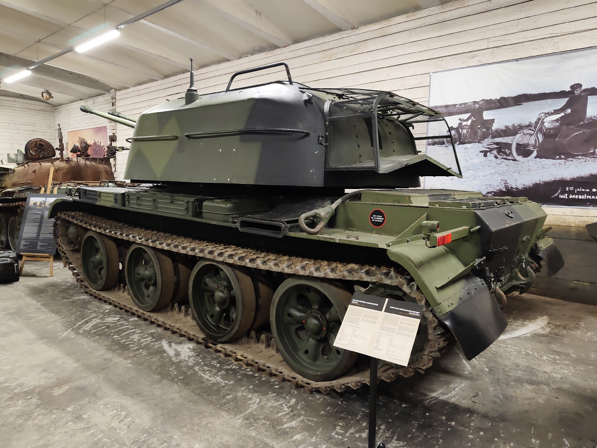 This image shows a military vehicle, likely a self-propelled anti-aircraft gun, displayed indoors in a museum. The vehicle is painted in a camouflage pattern of dark green and black. It has a tracked chassis with multiple road wheels and large metal tracks. The vehicle's turret is prominent, featuring a gun barrel protruding from the front and a cage-like structure on top. There are also signs or informational placards placed near the vehicle, one of which has text in a non-Latin script. In the background, other military vehicles are visible, as well as a large black and white photograph mounted on the wall depicting people on motorcycles. The environment appears to be a museum or exhibition hall with exposed ceiling beams and fluorescent lighting. This scene is likely located at the Viimsi Open Air Museum in Viimsi, Estonia, which is known for its collection of military vehicles and artifacts.
