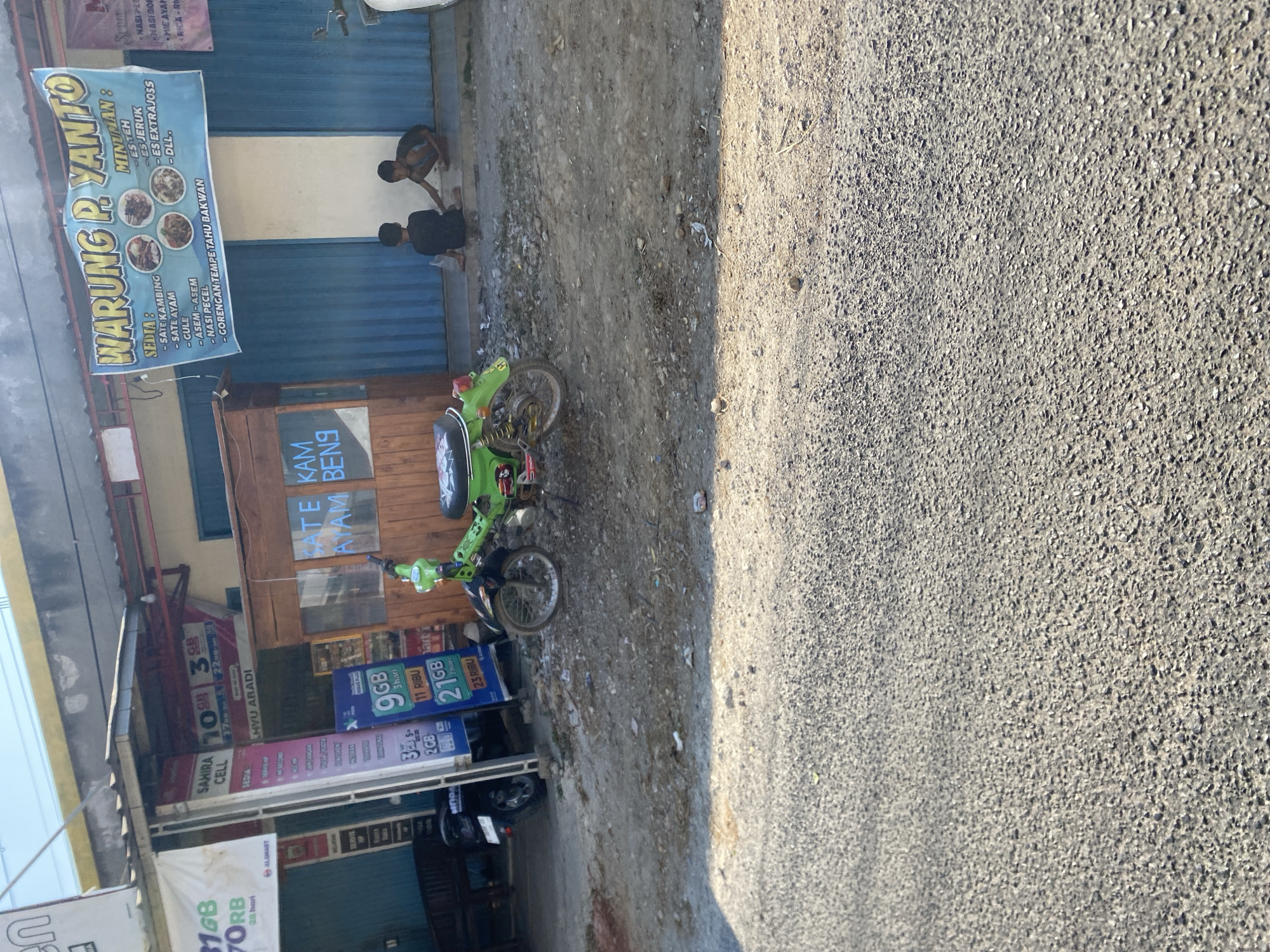 In a sunlit outdoor environment, a vibrant green motorcycle is parked on a gravelly area in front of a building with a blue corrugated metal facade. Two young boys are seated on the ground near the building entrance, interacting with each other. To the left of the motorcycle, a small wooden kiosk is visible. Above the kiosk, a blue banner advertising "WARUNG P. YANTO" with food and drink offerings is displayed. To the left of the kiosk, a stall with a white banner displaying "SAHIRA CELL" and mobile data packages is present, with promotional posters and items visible. In the background, the building has a light yellow overhang. The ground is covered in a mixture of gravel, dirt, and scattered debris.