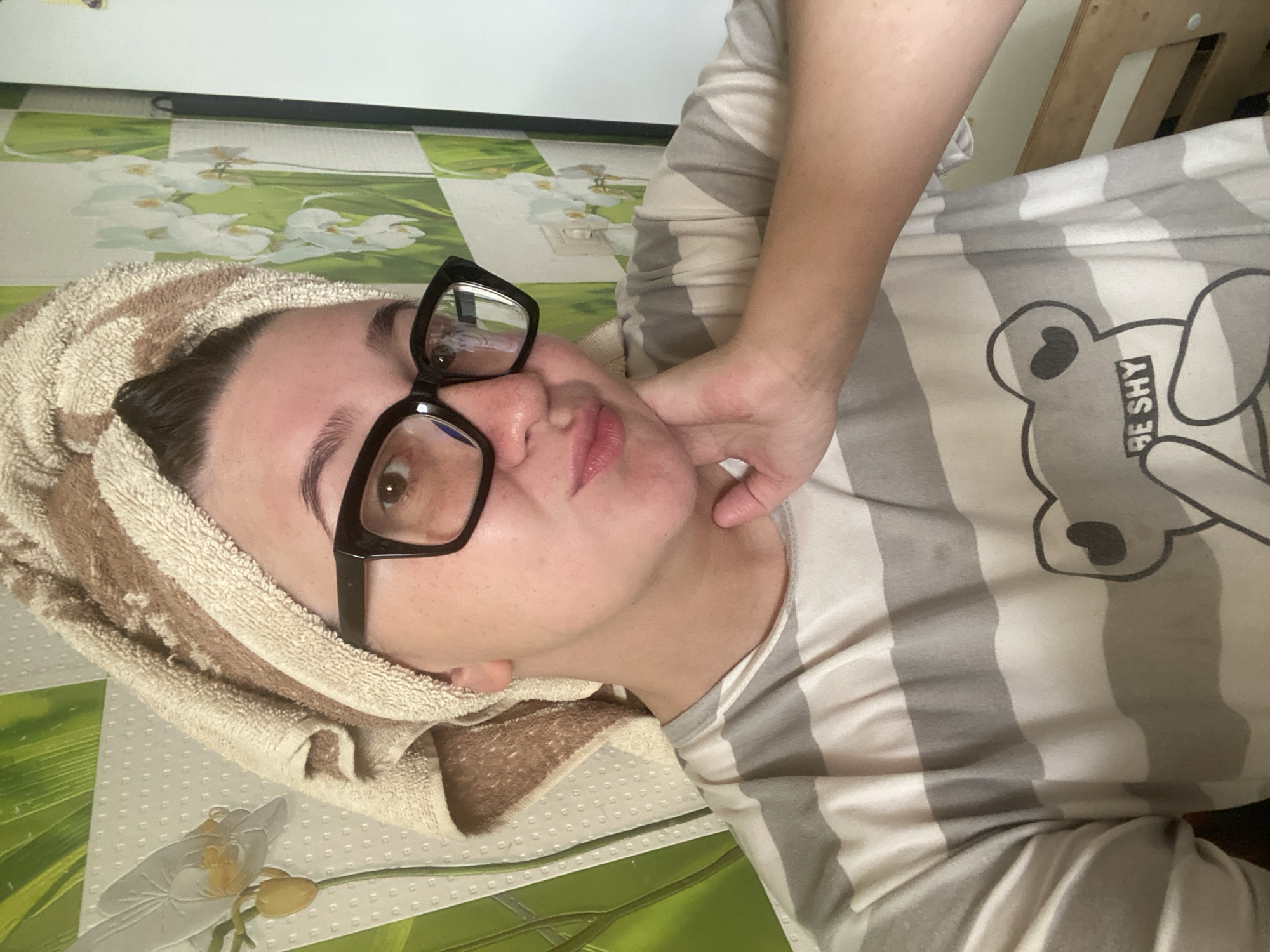 A person is depicted in a close-up, waist-up portrait captured in Chaikovsky, Russia. The individual, appearing to be female, is positioned centrally, looking directly into the camera with a slight smirk or puckered lip expression. They wear a light-colored, possibly beige and brown, terry cloth towel wrapped around their head. Large, black-framed rectangular glasses are worn, with reflections visible on the lenses. The individual's right hand (viewer's left) is resting against their chin and jawline. They are wearing a short-sleeved t-shirt featuring horizontal stripes of alternating light grey/white and darker grey. A graphic of a bear outline with a heart on its chest and the text "BE SHY" is printed on the front of the t-shirt. The background consists of a wall covered with wallpaper featuring a pattern of green foliage and white orchid flowers. A white electrical outlet is visible on the wall behind the subject's left shoulder (viewer's right). To the far right, a vertical white surface and a portion of a wooden structure, possibly furniture, are visible.