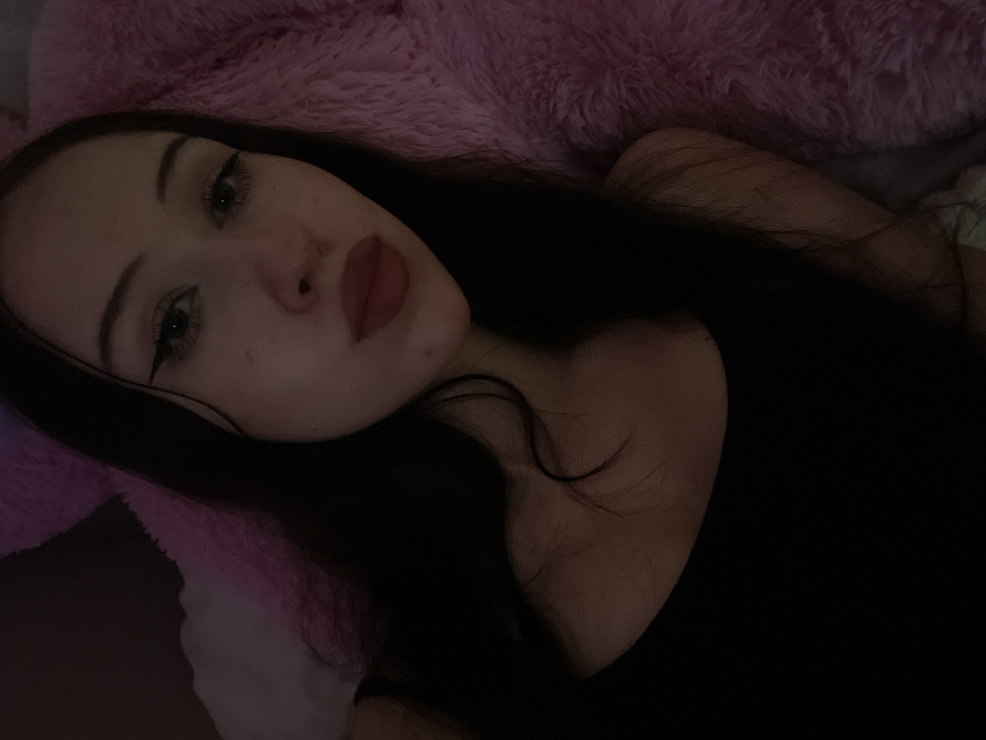 A young woman with long, dark hair and winged eyeliner is lying down on a fluffy pink surface, possibly a blanket or stuffed animal. She is wearing a black tank top. The lighting is dim, creating a soft, intimate atmosphere. The image appears to be a selfie, as the camera is angled towards the woman. No specific location in Votkinsk, Russia can be determined from the image itself.