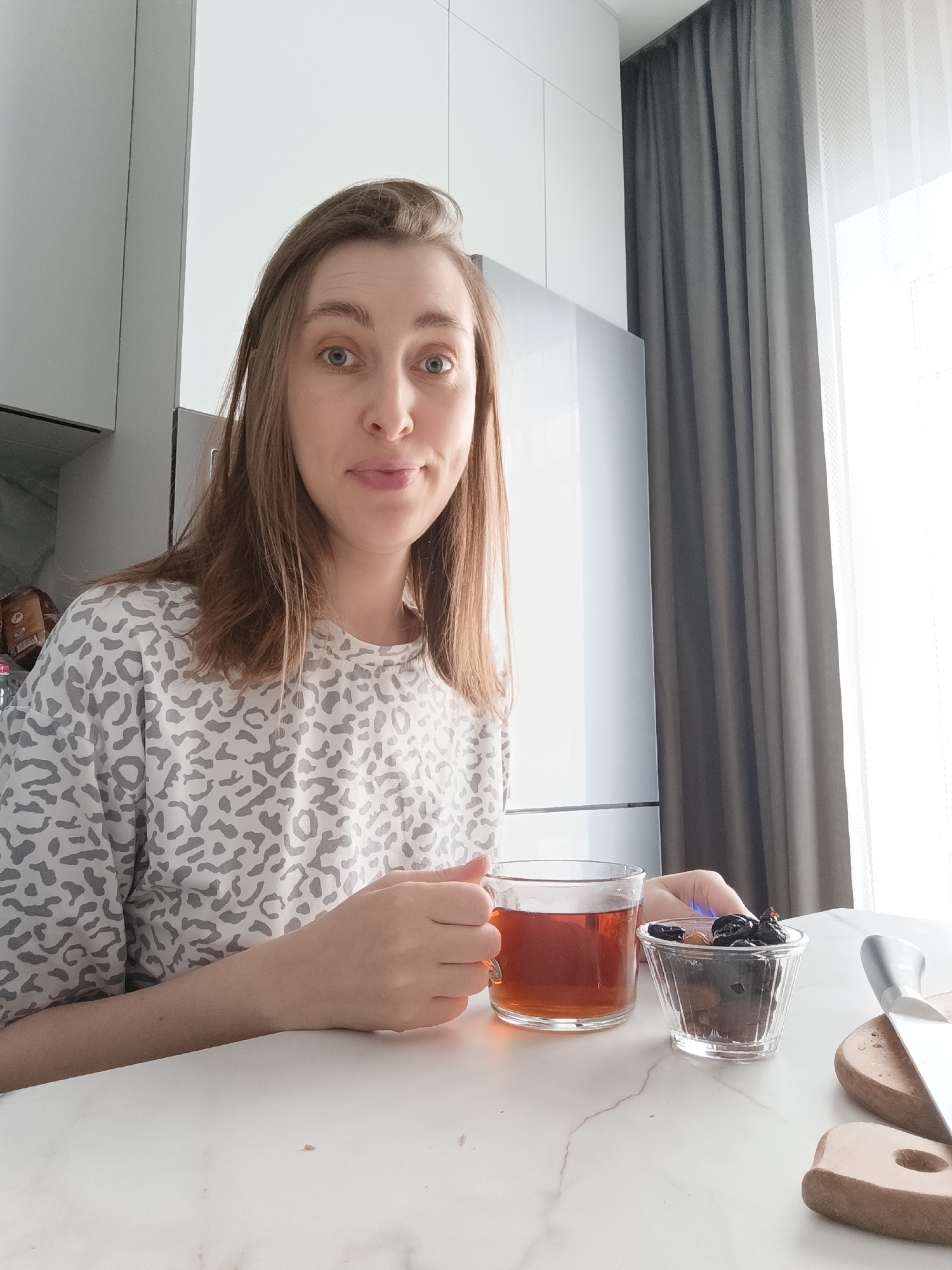 The image depicts a female individual in an indoor setting, identified as the city of Chaikovsky, Russia. She has medium-length brown hair and light skin, wearing a light-colored t-shirt with a grey leopard print pattern. Her eyes are directed towards the camera, and her lips are slightly pursed. She is seated at a white marble-patterned table, holding a clear glass mug containing a dark amber liquid, presumably tea, in her right hand. To her left, also on the table, is a small fluted glass bowl filled with dark dried fruits, likely prunes. Further to the right on the table, two small wooden cutting boards are visible, with a silver-bladed knife resting on one. The background features light grey and white kitchen cabinets on the left, a silver-colored refrigerator in the center, and a window with dark grey and sheer white curtains on the right, through which bright daylight is visible.