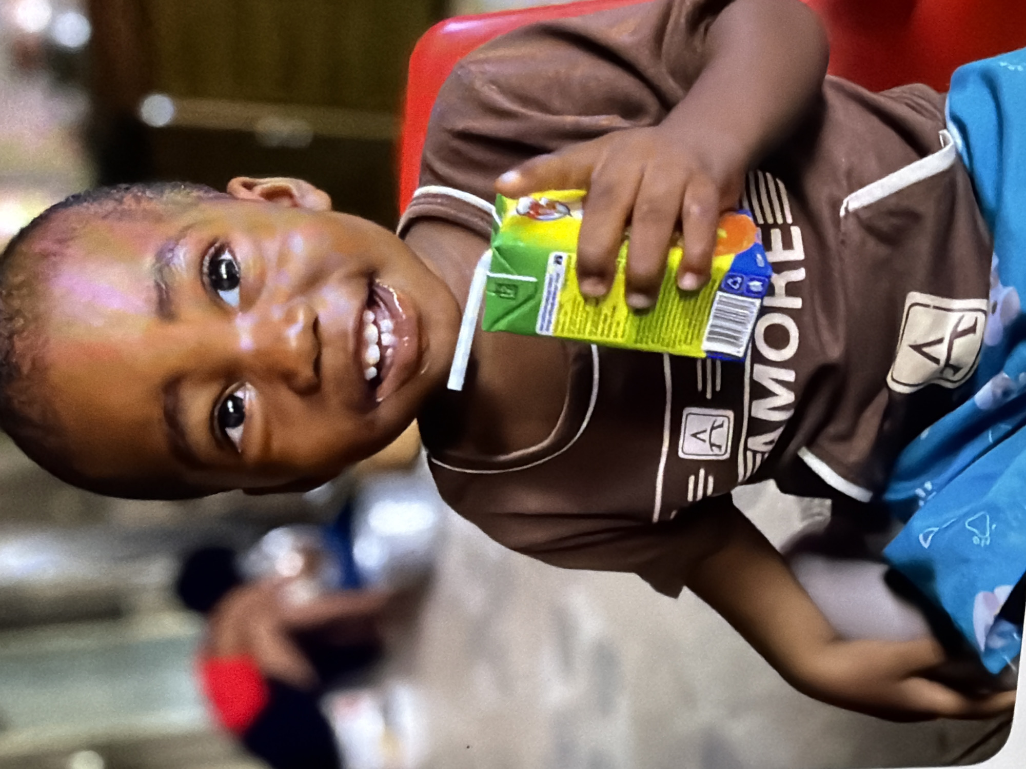 A child sits in a red chair, holding a juice box with a straw. The child wears a brown t-shirt with white trim and blue patterned shorts. In the blurred background, another person is visible, wearing a red and black garment. The setting appears to be indoors.