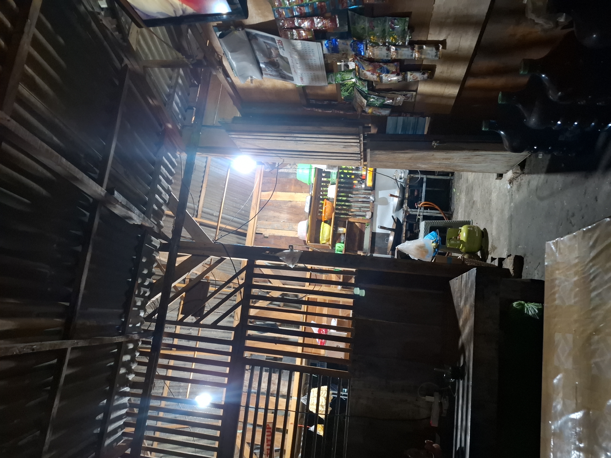 The image depicts the interior of a small, dimly lit eatery or shop, likely in Bulubrangsi, Indonesia, given the context. The structure appears rustic, with a corrugated metal roof supported by wooden beams and slats. The walls are also constructed of wood. In the foreground, two wooden tables are visible, the one closer to the viewer covered with a translucent plastic sheet.
The midground reveals a cooking area with what appears to be a stove and cookware, including woks. A green gas cylinder is prominently placed near the cooking area. Shelving units are stacked with various items, including containers, bowls, and what seem to be packaged goods. A calendar for 2026 is hanging on the wooden wall, along with an assortment of small packaged items, possibly snacks or condiments, displayed on the wall. Several large, dark-colored water jugs are lined up on the floor in the right side of the frame. The lighting is sparse, with a few bright bulbs illuminating sections of the space, creating areas of light and shadow. The overall impression is of a functional, working space, likely a local business.