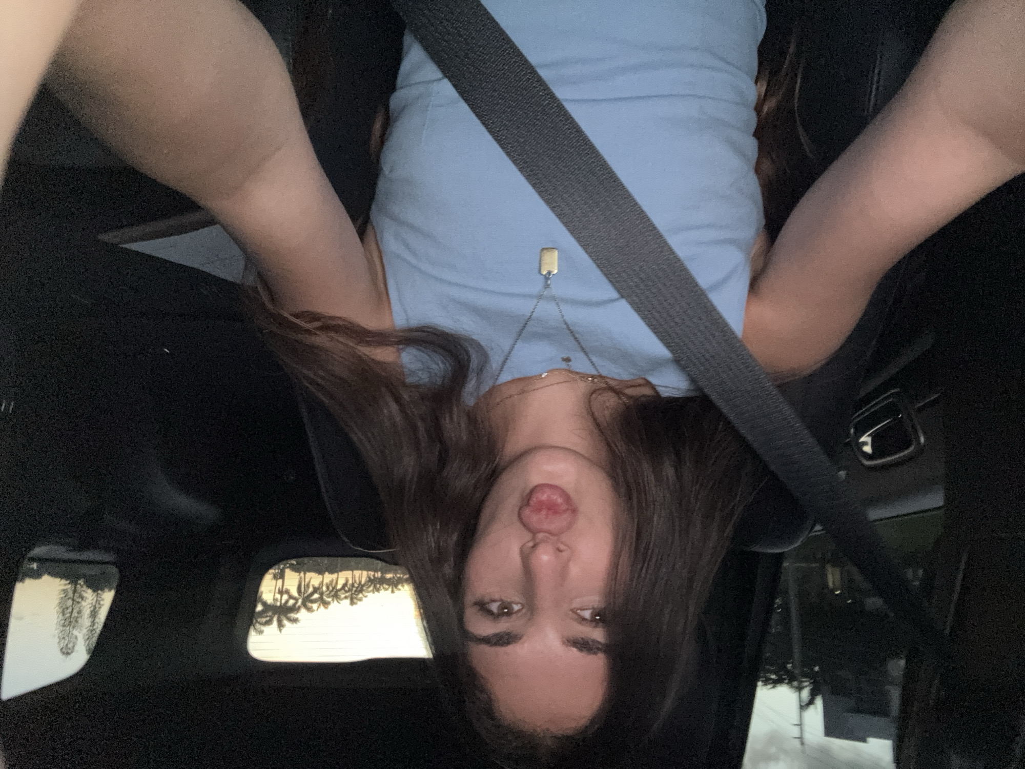 A young woman with long, dark brown hair and light brown eyes is seated in the front passenger seat of a vehicle in Boca Raton, United States of America. She is wearing a light blue sleeveless top and a silver necklace with two pendants, one small and circular, the other a longer, rectangular gold tag. Her lips are pursed in a "kiss" gesture, and she is looking directly at the camera, holding it herself for a selfie. A dark grey seatbelt crosses her chest. Through the driver's side window on the left, an overcast sky is visible above dark building structures and trees, with a utility pole and power lines also discernible. Through the rear windows, a clear sky with a sunset gradient (orange/yellow near the horizon) is visible, silhouetting a distinct row of numerous palm trees on the left and other trees on the right. The overall lighting suggests dusk. The car's interior appears dark, with a door handle visible on the left side.