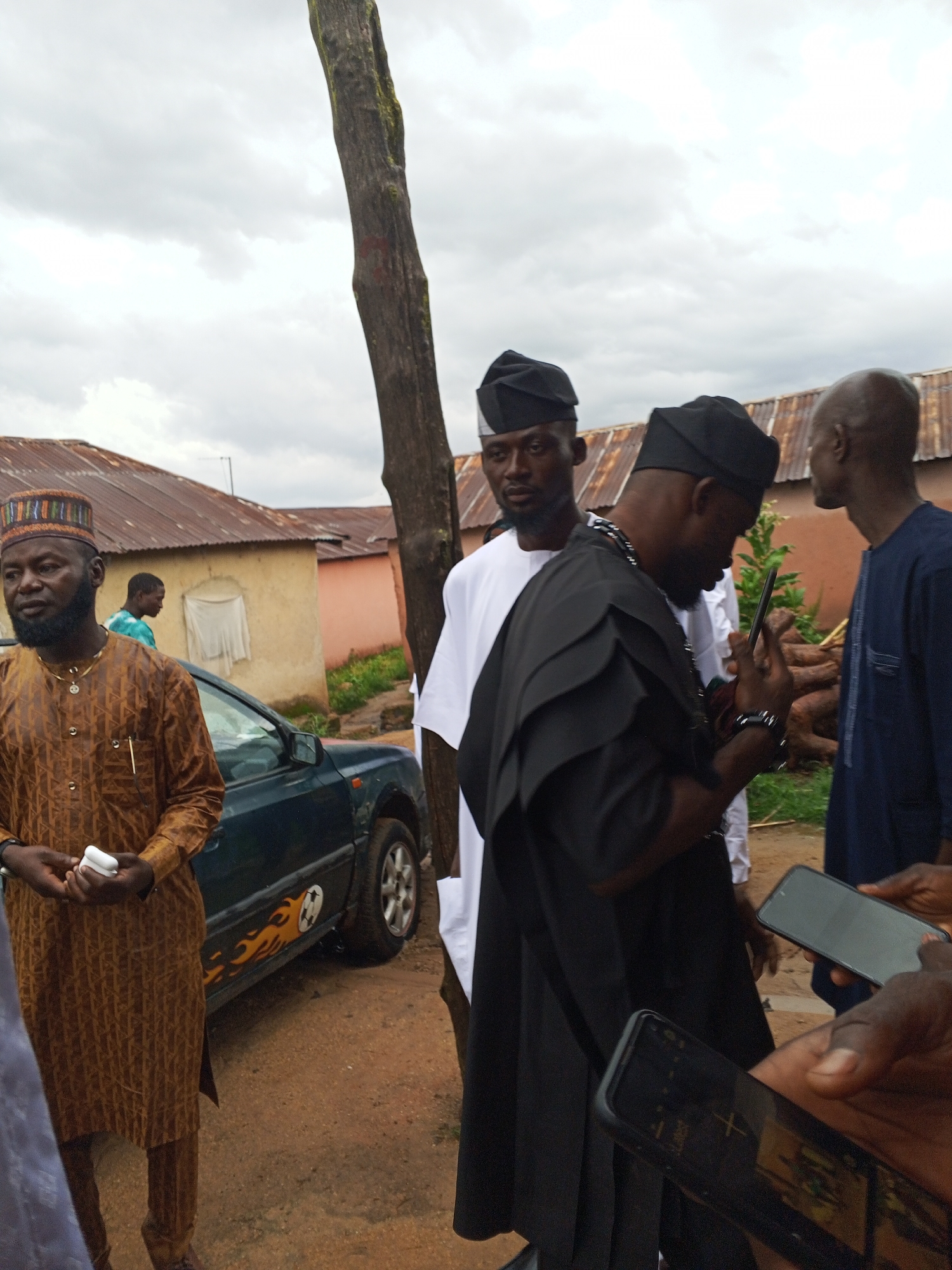 In Panyam, Nigeria, under an overcast sky, a group of men in traditional attire gathers outdoors. The scene is set against a backdrop of residential buildings with corrugated metal roofs. In the foreground, a man wearing a patterned brown tunic and a decorative cap holds a white object in his hands. To his right, another man, dressed in a black robe with layered, structured sleeves and a matching black cap, is looking down at a smartphone he holds. To the left of him, a man in a white robe and black cap is partially visible. Further back, another man in a dark blue tunic is seen. A dark green car with a flame decal is parked behind the man in the brown tunic, and a large tree trunk stands prominently between the man in the black robe and the man in the blue tunic. The ground is covered in reddish-brown dirt.
