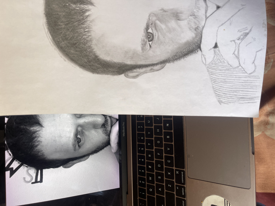 The image displays a MacBook Pro laptop with its screen showing a black-and-white photographic portrait of a man. The portrait is cropped to the right half of the man's face (viewer's left), featuring dark hair, a beard, and his right hand partially covering his chin. His right eye (viewer's left) looks directly forward. Visible on a white wall in the background of the displayed photograph is the word "JOS", indicating the photograph's origin in Jos, Nigeria. A sheet of white paper, containing a pencil sketch of the same man in a similar pose, partially covers the right side of the laptop screen and keyboard. The sketch is rendered in pencil, showing the man's right facial features and hand with visible pencil lines and shading. The laptop and sketch-bearing paper rest on a patterned surface, appearing to be textile, with shades of brown and red.