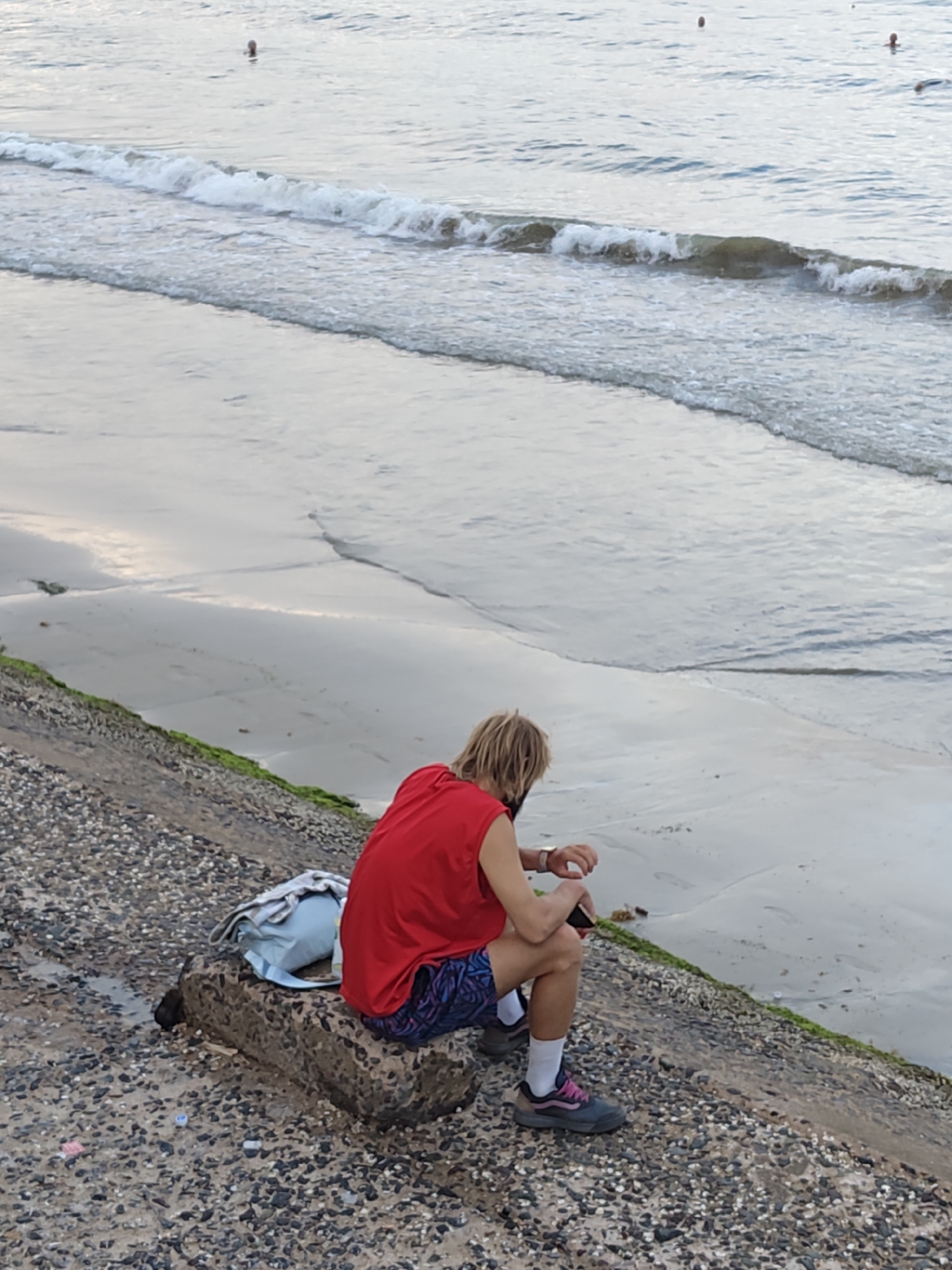 A person with blonde hair, wearing a red tank top and patterned shorts, sits on a large rock on a beach. The person is facing the ocean, with their head slightly bowed and their hands clasped in their lap. A light blue backpack rests on the rock next to them. The person appears to be looking at their hand, perhaps a watch or a phone. The beach is made of pebbles and has some green algae growing on the rocks. Gentle waves are rolling in from the sea. In the distance, a few other people can be seen in the water. The sky is overcast, and the lighting suggests it might be early morning or late afternoon. The location is identified as Nha Trang, Vietnam.