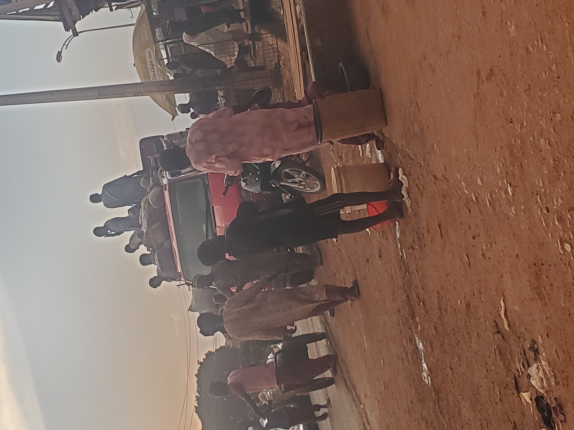 The image depicts a crowded street scene, likely in the city of Funtua, Nigeria, during what appears to be either early morning or late afternoon due to the soft lighting. In the foreground, several individuals are visible. A person in a pink garment is standing next to a large, light-colored barrel, with another barrel and a dark basin nearby. To their left, a young person in a black shirt and shorts stands with their feet in what seems to be shallow water or mud. Further left, a person in a pink shirt and dark pants is pushing a cart with two wheels. Behind them, a person in a tan garment walks away from the camera, and another person in dark clothing is also present.
Dominating the middle ground is a red truck, heavily laden with goods, possibly bags of produce or other merchandise. Several people are perched on top of the truck, some appearing to be communicating or observing their surroundings. One person is speaking on a mobile phone. Another individual, dressed in blue, is seated on the truck.
In the background, a yellow umbrella, advertising "Glo Café app" and "VOICE," is visible, suggesting a commercial area. A street light and the side of a building are also present. The ground is unpaved and appears to be dirt or compacted earth, with some scattered debris and puddles. The overall impression is one of a bustling, active street in a Nigerian urban environment.