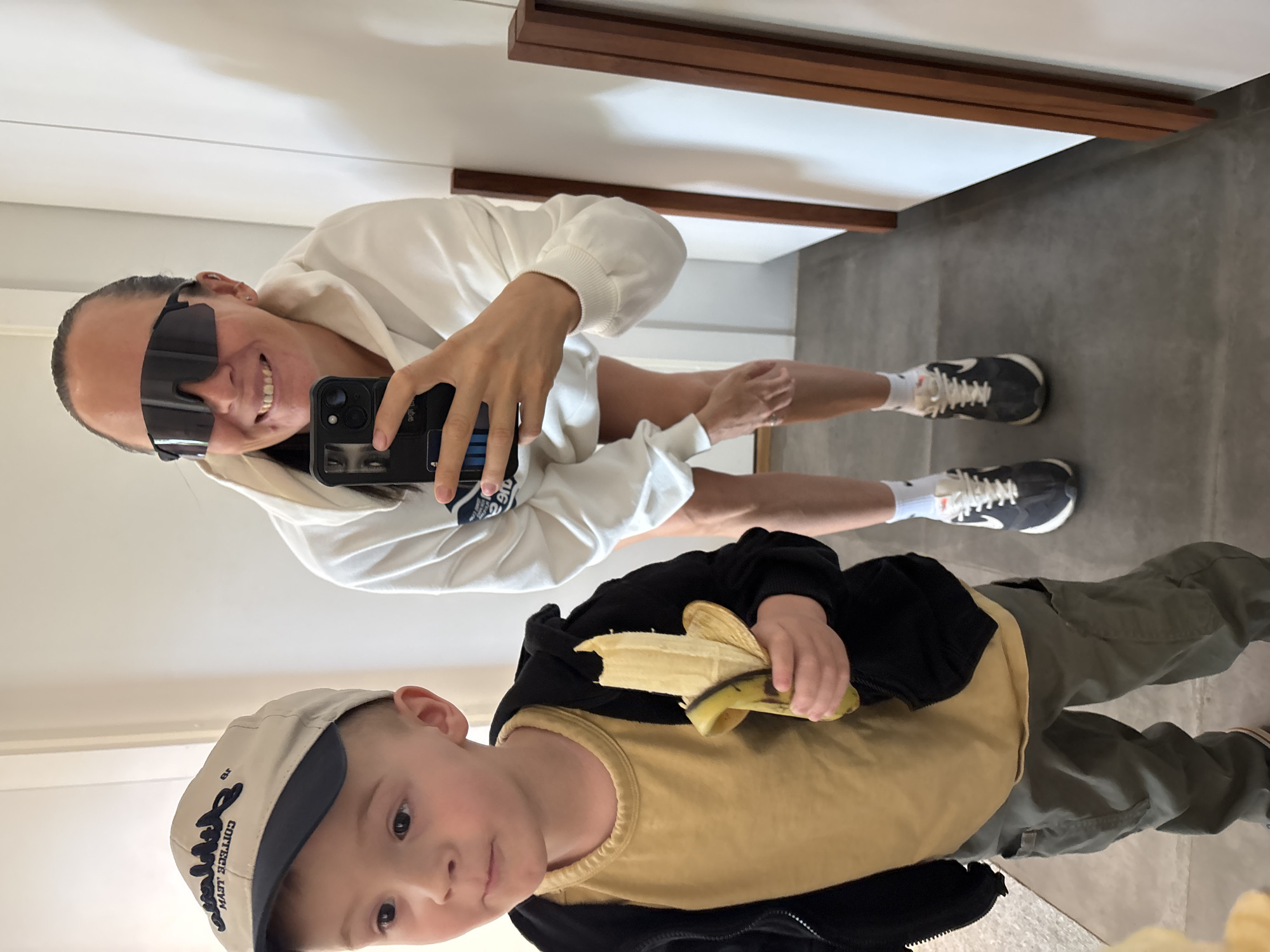 A woman wearing a white hoodie and dark sunglasses takes a selfie in a mirror. She is wearing gray and white sneakers. A young boy stands in front of her, holding a banana. He wears a light brown cap, a black hoodie, an ochre t-shirt, and green pants. They are standing in a room with a gray floor and white walls. The location is unknown.
