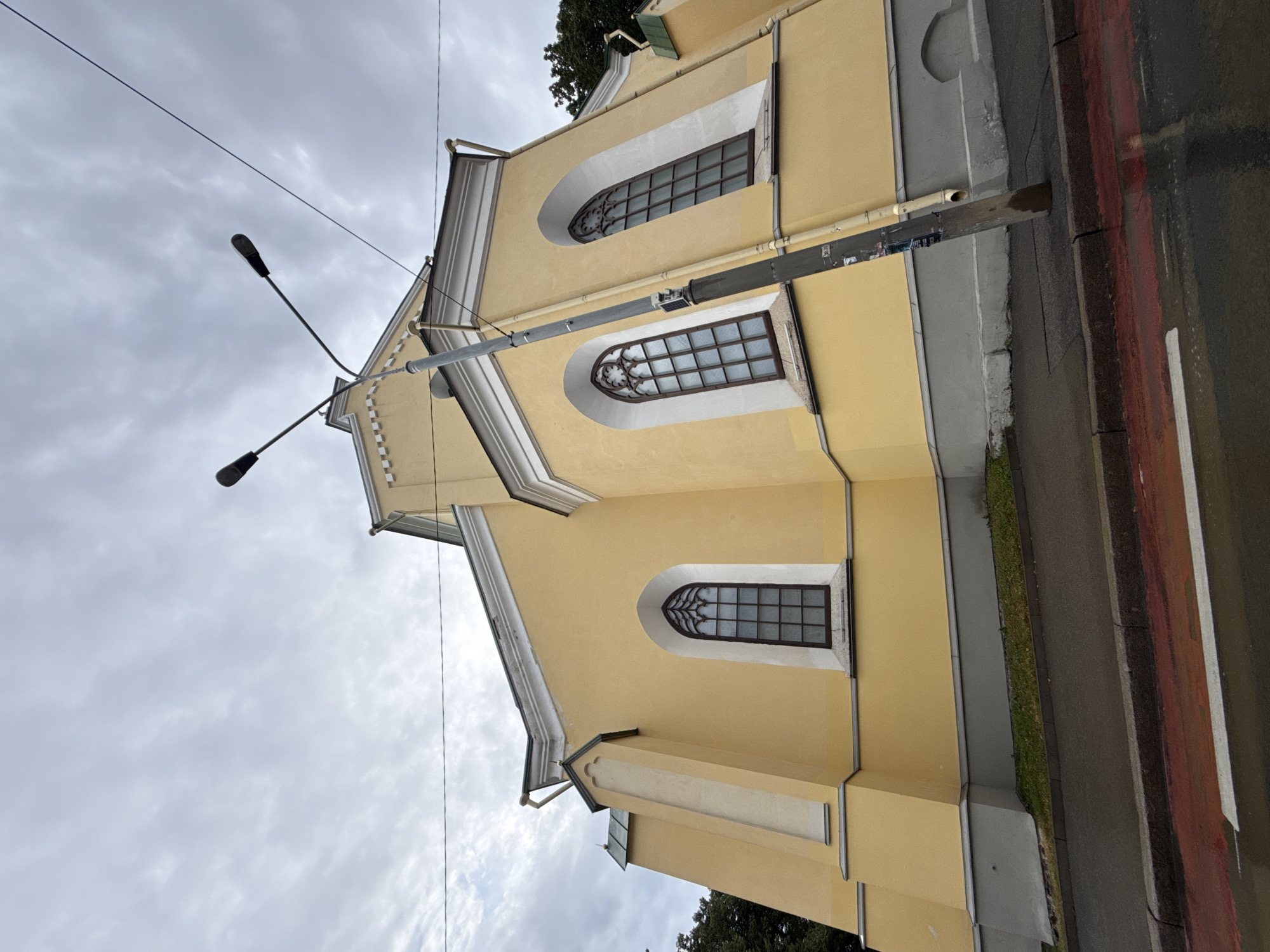 The image captures a low-angle exterior view of the yellow facade of St. John's Church in Tallinn, Estonia. The overcast sky creates a muted light. A tall, grey lamppost with two lights stands prominently in the foreground, angled towards the church. The church features tall, arched windows with intricate dark frames. The building's base is a grey plinth, and a patch of green grass and a dark asphalt road with red and white markings are visible in the lower portion of the frame. The road appears wet, suggesting recent rain. Power lines are visible crisscrossing the sky.