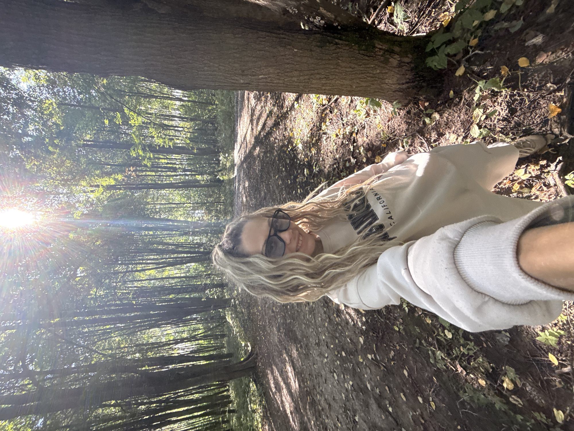 A woman is taking a selfie in a forest. She has long blonde hair pulled into a ponytail and is wearing sunglasses and a light-colored sweatshirt. The sweatshirt has "California" written on it in black letters. She is smiling at the camera. The forest floor is covered in dirt and fallen leaves, and sunlight streams through the trees. The location is the city of Rusanovo, Russia.