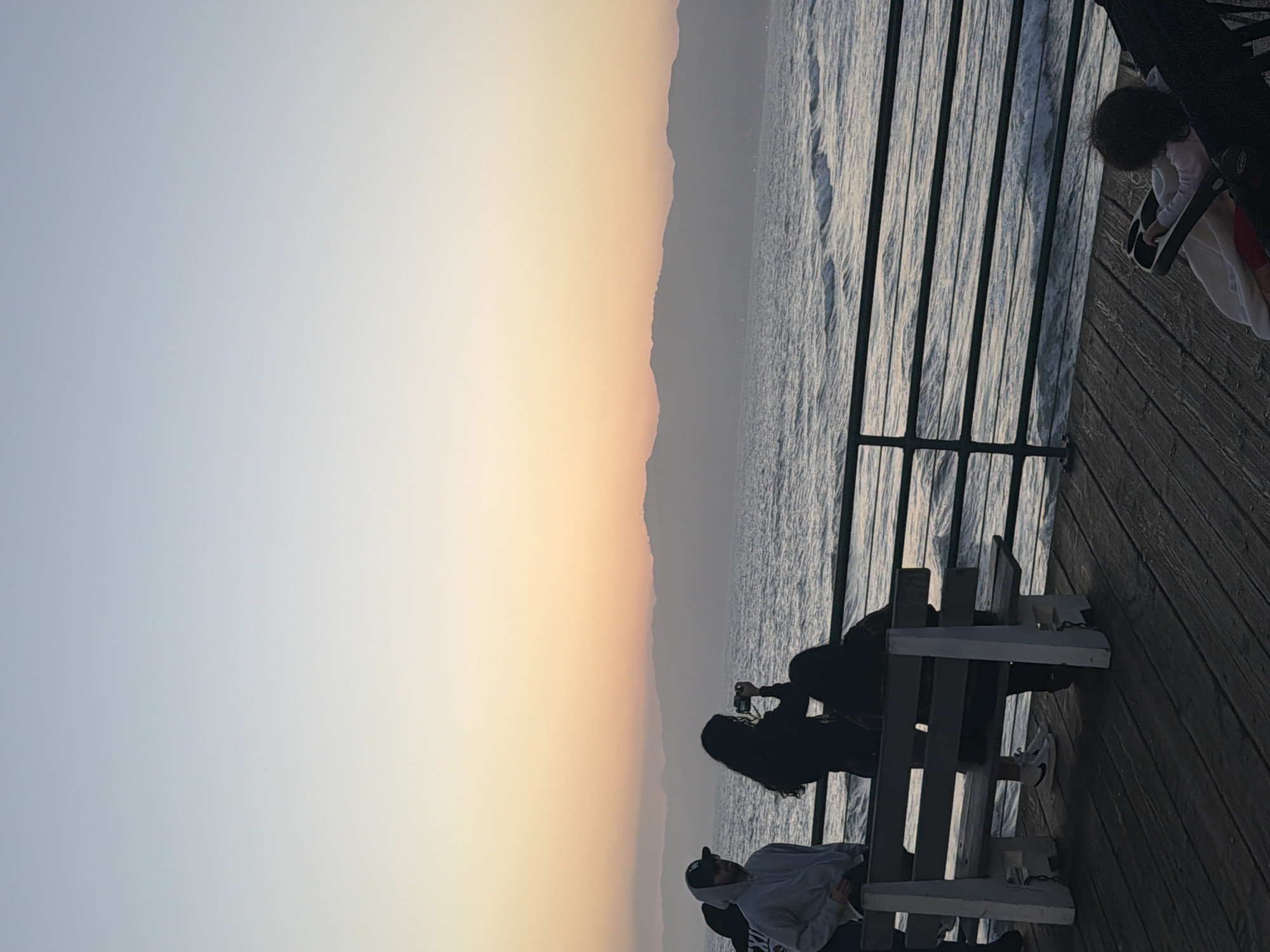 The image captures a serene scene at Santa Monica Pier in California, United States. The sky displays a gradient of colors, transitioning from light blue to a warm orange near the horizon, indicative of either sunrise or sunset. A range of mountains can be observed in the middleground, with the vast expanse of the ocean stretching out to meet them.
On the wooden pier, a group of people are sitting on a bench. One individual is holding a camera, likely capturing the scenic view. Further along the pier, a child is visible in a stroller, suggesting a family outing. The pier's railings run parallel to the shoreline, adding depth to the composition. The wooden planks of the pier are visible, contributing to the structural texture of the scene. The overall atmosphere is one of relaxation and enjoyment of the natural surroundings.