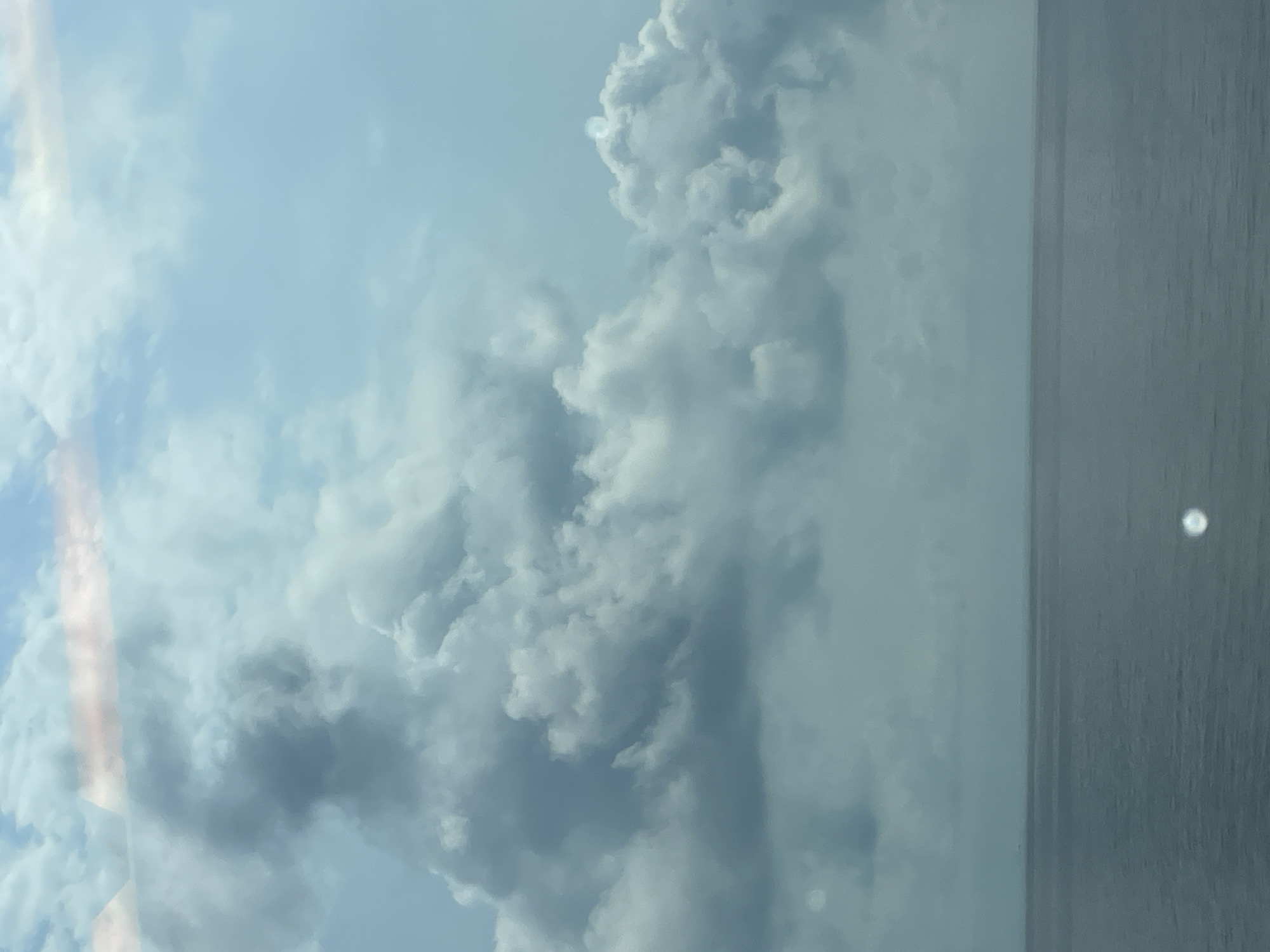 The image shows a vast expanse of calm, dark blue ocean meeting a hazy, overcast sky. A thin line of distant land is visible on the horizon. The sky is filled with heavy, grey clouds, with lighter patches of white and a faint pinkish hue at the top, suggesting a possibly overcast sunrise or sunset. The overall atmosphere is subdued and muted, with no signs of human activity or specific landmarks from Sochi, Russia, visible. The image appears to be taken from a watercraft or a high vantage point overlooking the sea.