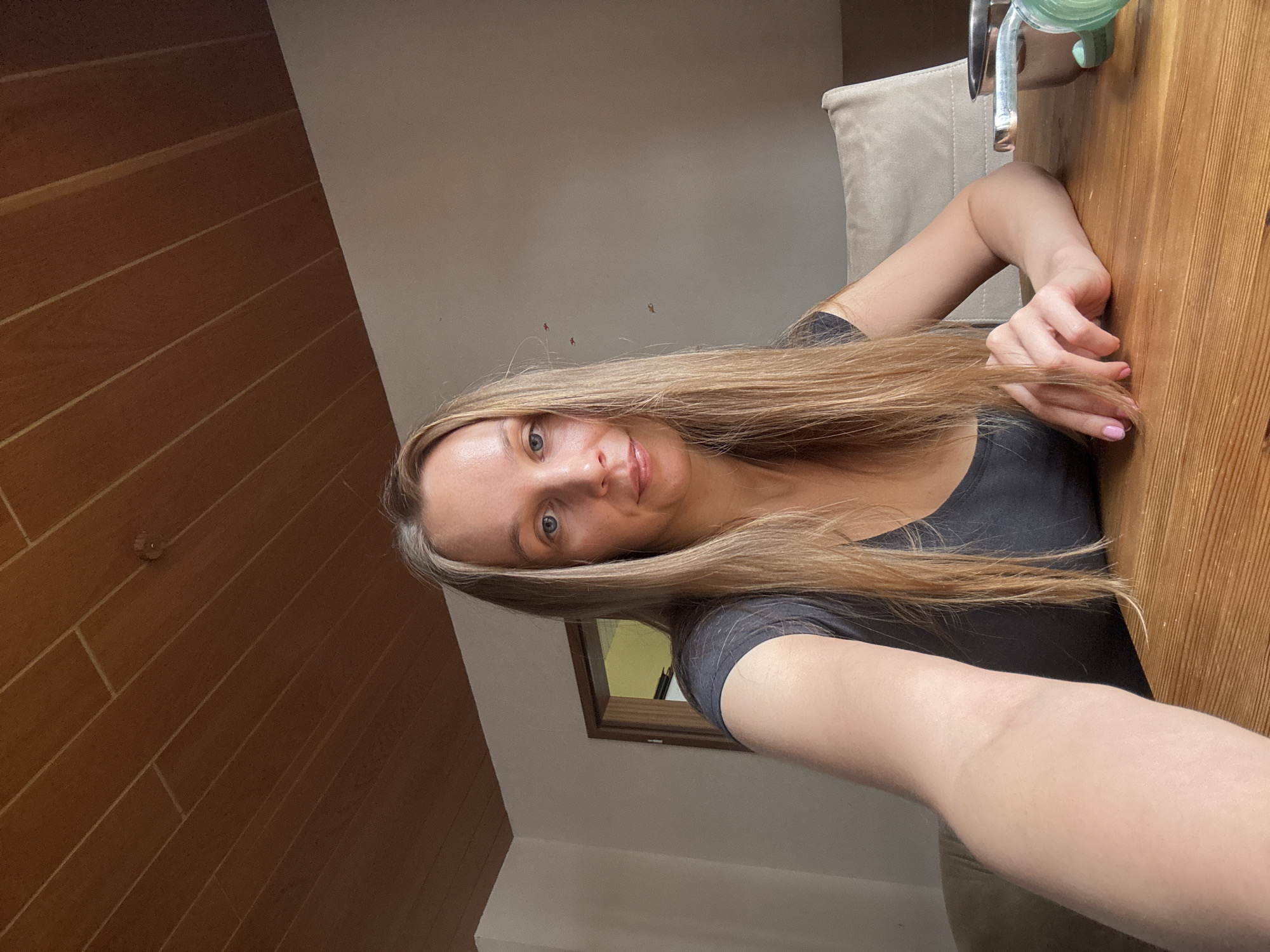 A woman with long blonde hair is sitting at a wooden table, taking a selfie. She is wearing a dark grey t-shirt. In the background, there is a doorway leading to another room, and a chair with a beige cushion. The ceiling is made of wood panels. A metal bowl and some green object are on the table to the right of the woman. The scene appears to be indoors, possibly in a home. The image does not provide enough information to determine a specific location within Überherrn, Germany.