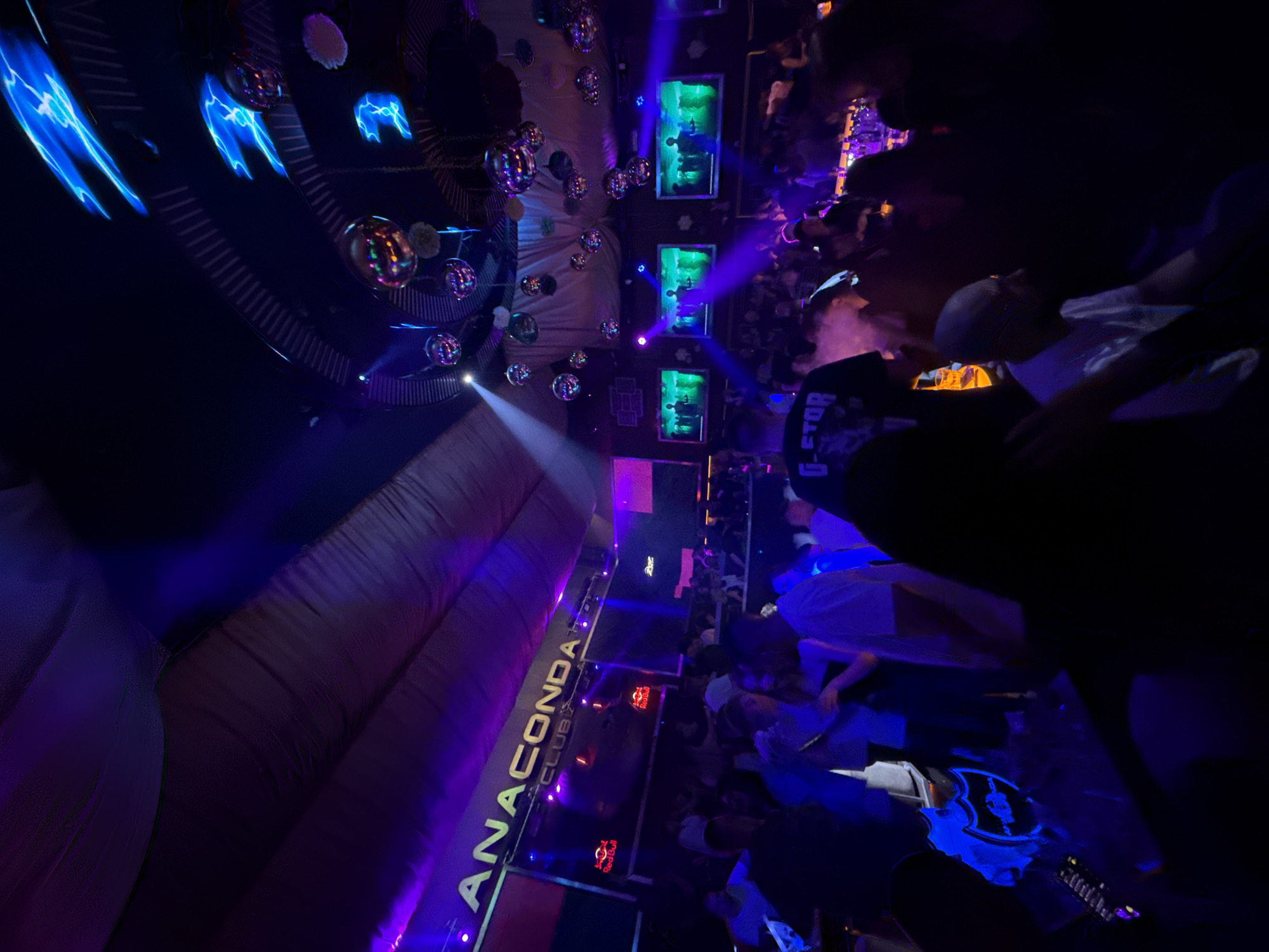 The image depicts the interior of Anaconda Club, located in Pleven, Bulgaria. The nightclub is dimly lit with blue and purple lighting effects. The ceiling is adorned with disco balls and projected light designs. Multiple screens display images. A crowd of people is visible, with individuals standing and interacting.