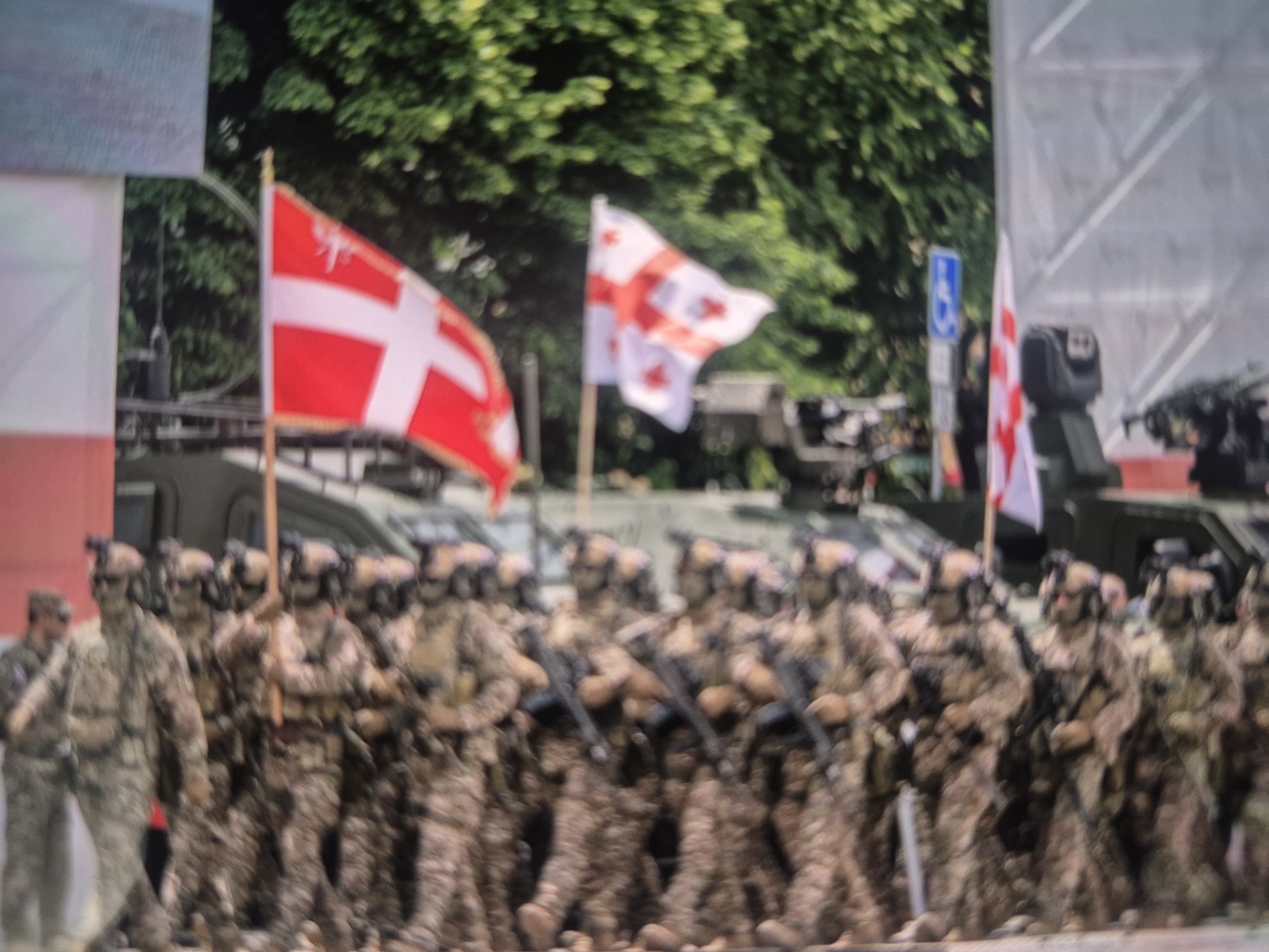 In Tbilisi, Georgia, a military parade features soldiers in camouflage uniforms carrying weapons. Behind them are military vehicles. Three flags are visible: a red and white cross flag, the Georgian flag, and another Georgian flag. Trees are in the background. A blue sign is also visible behind the flags.