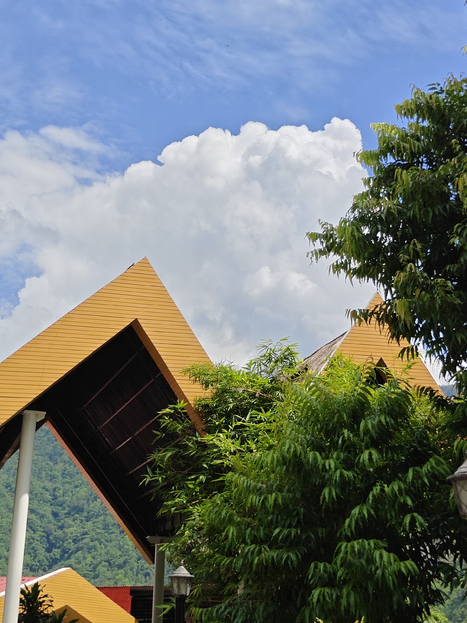 The image depicts an outdoor scene under a bright sky with white clouds. In the foreground, a modern building with a distinctive golden, angular roof structure is visible. The roof has horizontal paneling and an exposed dark-colored interior framing. To the right of the building, lush green foliage from trees, including bamboo, obstructs the view. In the background, a verdant, tree-covered mountainside is visible through the open architecture of the building. The overall impression is one of a scenic location integrated with modern architecture, likely in a tropical or subtropical environment. There are no people or animals in the frame. The scene is consistent with the landscape of Ichchhakamana, Nepal, known for its natural beauty and mountainous terrain.