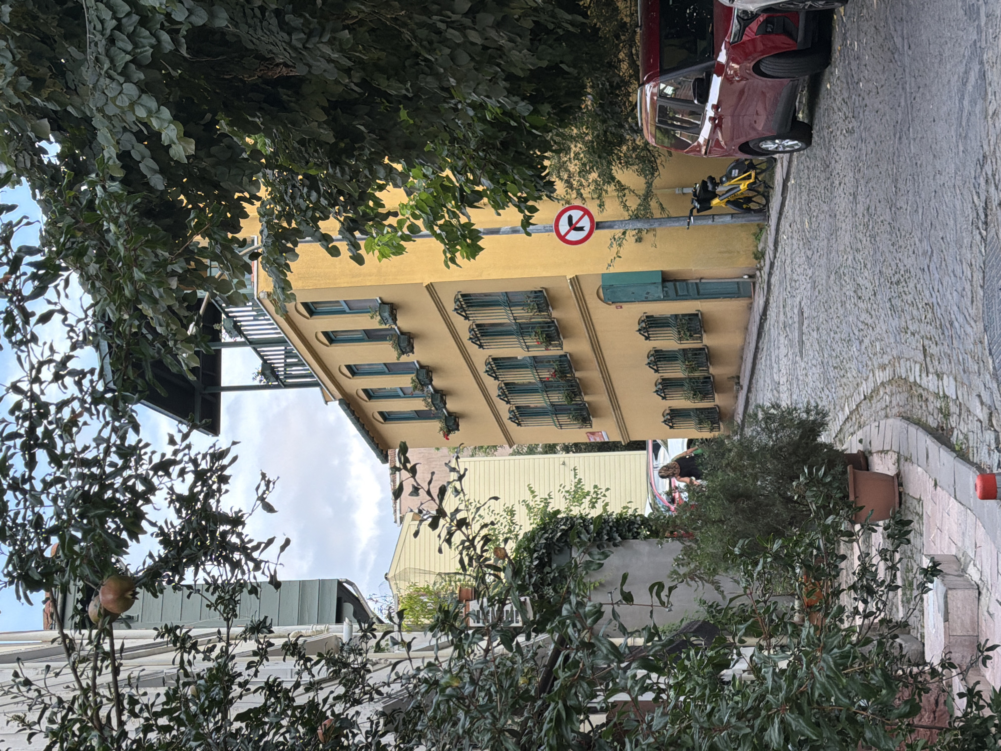 This image captures a narrow, cobblestone street in Üsküdar, Turkey, likely in the Kuzguncuk neighborhood, known for its charming, old-world architecture. The street is lined with buildings of varying heights and colors. On the right, a vibrant yellow building with green-shuttered windows and decorative iron railings dominates the view. To its left, a portion of a lighter-colored building with similar architectural features is visible. The street is partially shaded by lush green trees, their branches laden with what appear to be pomegranates, particularly in the upper left quadrant of the image.
Several objects are present on the street. A bright yellow bicycle with black accents is parked near the yellow building. A red SUV is parked on the right side of the street, partially obscuring another vehicle behind it. A traffic sign indicating "no right turn" is posted on a pole beside the yellow building.
In the middle ground, a woman with dark hair wearing a dark top and lighter bottoms is walking away from the camera, carrying a green bag. Potted plants and greenery are visible along the sides of the street and on the buildings, adding to the picturesque atmosphere. The sky is overcast, suggesting a cloudy day. The overall scene depicts a tranquil, residential street with a strong sense of local character.
