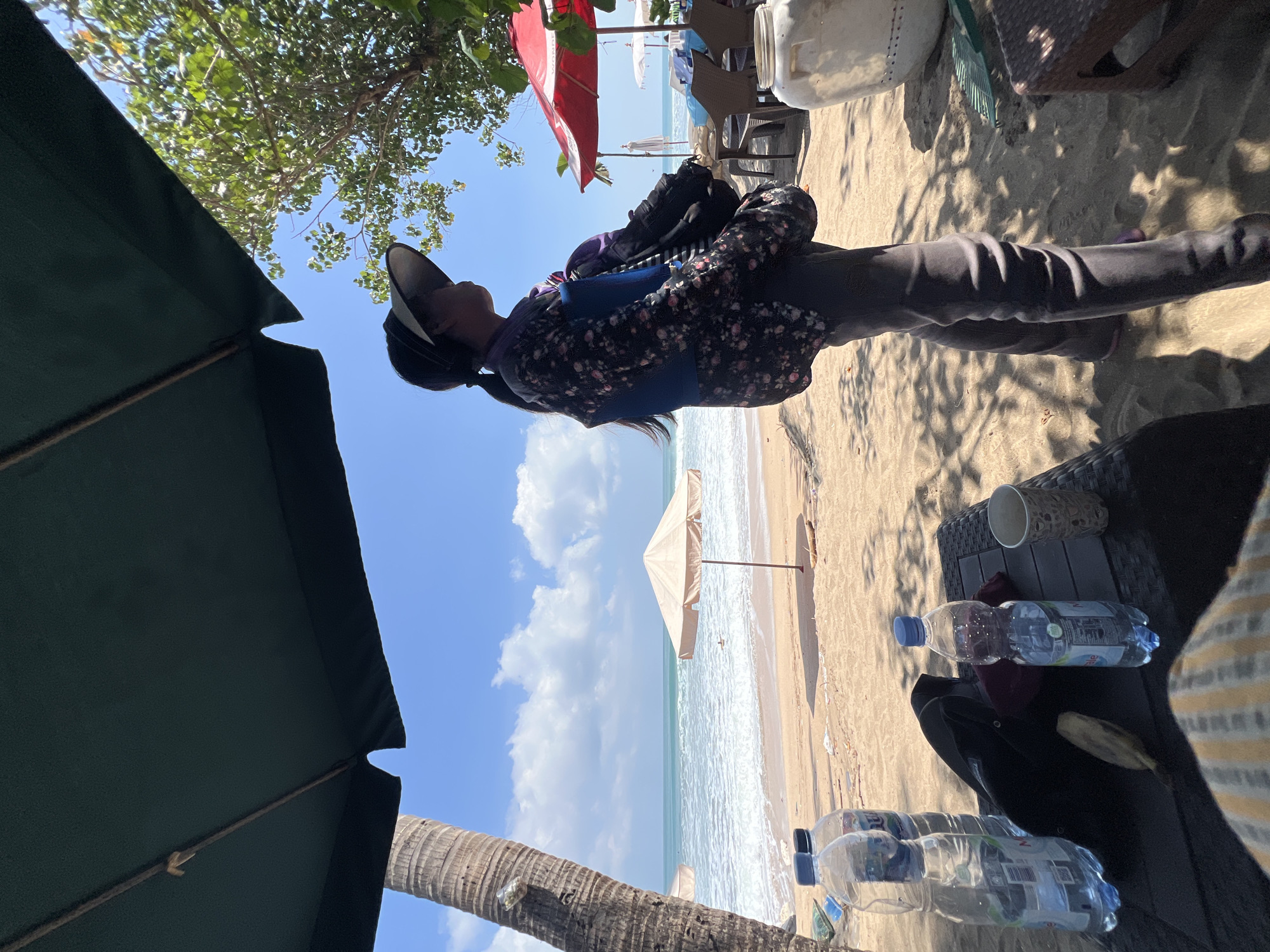 A person stands on a sandy beach in Kuta, Indonesia, under the shade of a large dark green umbrella. The person, facing right, wears a dark floral print long-sleeved shirt, a blue vest, dark pants, and a wide-brimmed hat. A ponytail is visible. Several plastic water bottles and a banana lie on a dark woven table in the foreground. In the background, the ocean waves crash onto the shore under a bright blue sky with scattered white clouds. Two beach umbrellas, one beige and one red, are visible further down the beach. Trees line the edge of the sand, casting dappled shadows on the ground. The general atmosphere is that of a sunny day at a tropical beach resort.