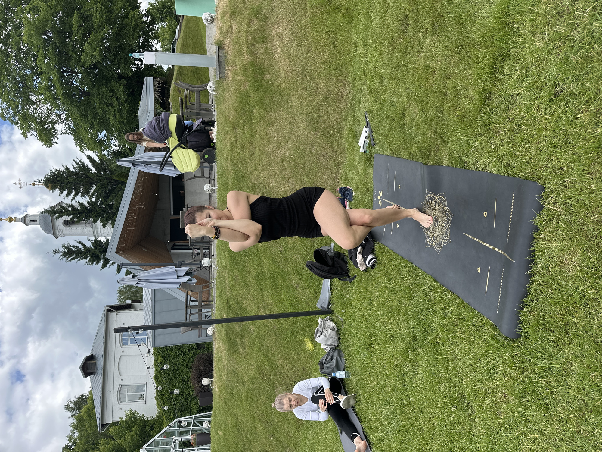 An outdoor scene captures a group of people engaged in what appears to be a yoga or fitness session on a bright, overcast day in Vladimir, Russia.
The central figure is a woman in a black sleeveless top and shorts, performing a balancing pose akin to a modified Garudasana (Eagle Pose) on a dark grey yoga mat. Her left leg is wrapped around her right, and her arms are crossed, with her hands resting on her shoulders. The mat features a prominent gold mandala design and the visible brand name "LUNU" in gold script, along with other geometric markings. Her bare feet are on the mat.
To her right, another woman with blonde hair, dressed in a white long-sleeved top and dark leggings, is seated on a separate dark yoga mat, holding a cup. Nearby, a water bottle and a black bag are placed on the lush green grass. Further in the background, a third person stands near a distinct lime-green baby stroller.
The setting is a well-maintained grassy area, possibly a park or outdoor facility. Several buildings are visible in the background: a modern structure with a wooden roof and outdoor seating under grey umbrellas, and a traditional white multi-story building with arched windows. Most notably, an Orthodox church with a white spire, golden dome, and cross stands prominently against the cloudy sky, providing a strong architectural clue to the location. Large green trees and decorative string lights add to the ambiance. The overall impression is one of a serene and focused outdoor activity.
