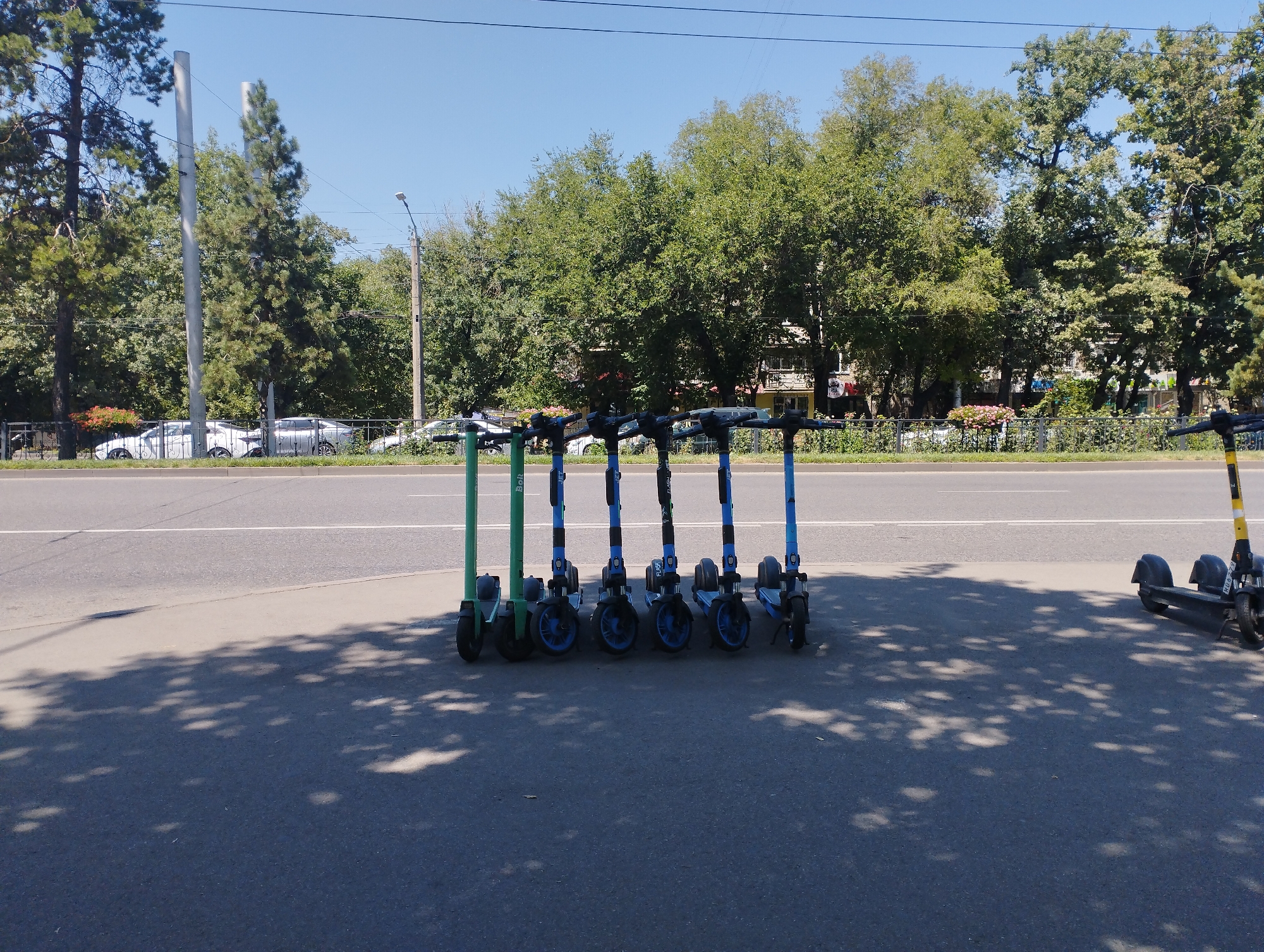 A row of electric scooters are parked on a sidewalk next to a road in Almaty, Kazakhstan. The scooters are a mix of green, blue, and black, with some displaying branding. In the background, there is a road with a white line marking, trees, and buildings. The sky is clear and blue. The image appears to be taken during the daytime.