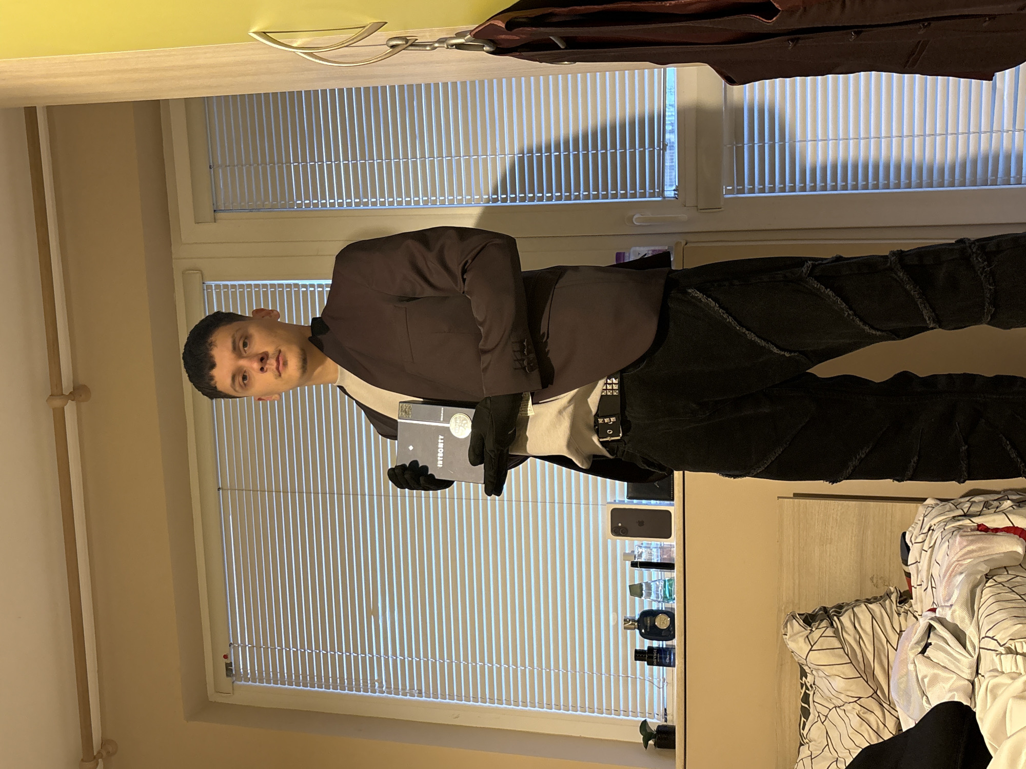 This is an indoor, full-body portrait of a young man, likely taken in Plovdiv, Bulgaria, who is posing directly for the camera. He appears to be in his late teens or early twenties, with short, dark hair and a neutral expression.
He is dressed in a dark brown or charcoal suit jacket over a lighter undershirt and dark gloves on both hands. His trousers are dark, possibly black jeans, featuring textured or distressed patterns, particularly on his left leg. He also wears a dark belt with a silver-colored buckle. In his left gloved hand, he holds a rectangular object, possibly a book or a case, with the word "INTEGRITY" clearly visible on its spine or cover.
The background features a large window with venetian blinds, through which natural daylight streams, creating vertical patterns of light and shadow on the wall. To the man's right, a surface, possibly a shelf or dresser, holds several items. These include a dark rectangular box, likely for an iPhone, and multiple bottles of cologne or perfume, one of which is identifiable as "Dior Sauvage." A small green potted plant is also visible on this surface. To the far right, part of a bed with white and black patterned bedding is partially visible. The walls are a light, neutral beige or cream color. The general setting suggests a bedroom or living area during the daytime.