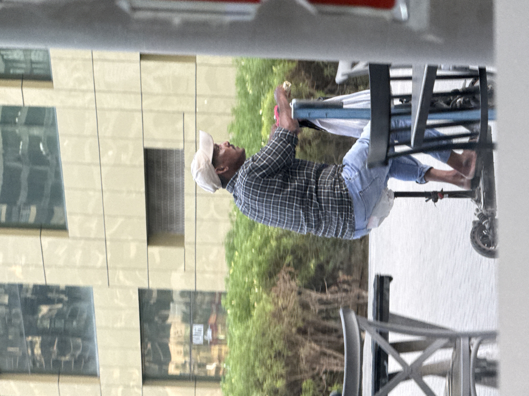 A man wearing a plaid shirt, blue jeans, and a light-colored baseball cap is riding an electric scooter. He is barefoot and appears to be holding something in his right hand. The scooter has a blue pole extending upwards. The scene appears to be outdoors, possibly on a sidewalk or promenade, with a modern building in the background featuring large windows reflecting the sky. Green shrubbery is visible behind the man. In the foreground, parts of outdoor furniture, specifically chairs and a table, are visible, suggesting a cafe or restaurant setting. The image is slightly motion-blurred, indicating movement. The architecture and vegetation suggest an urban environment in Dubai, United Arab Emirates.