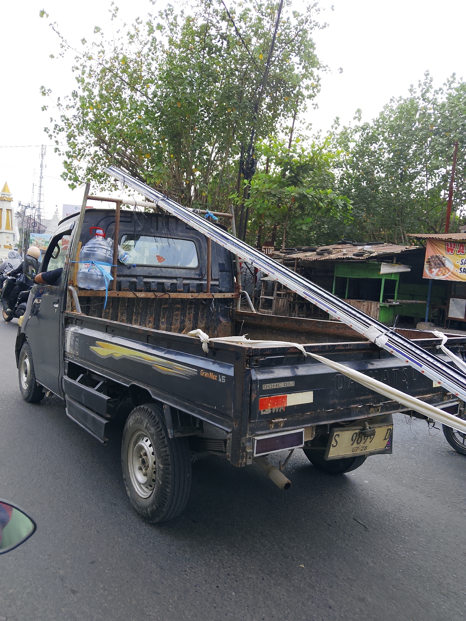 The image shows a black pickup truck with a large bottle of water secured in its cab, on a street in Balun, Indonesia. The truck is carrying long, cylindrical objects, possibly pipes or poles, extending from its bed and secured with rope. The truck's license plate reads "S 9099 D" with smaller text below. To the left of the truck, a person on a motorcycle wearing a helmet is visible. In the background, there are trees, a small building with a green facade and a corrugated metal roof, and a sign advertising food, possibly "SATE". Power lines and streetlights are also present. The street is paved and appears to be a typical urban environment. The sky is overcast.