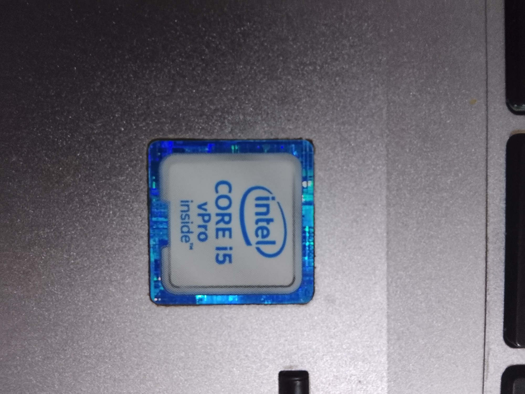 The image shows a close-up of an Intel Core i5 vPro sticker on a laptop. The sticker is blue and white. The laptop's surface is metallic gray. A portion of a keyboard, specifically the edge of several keys, is visible on the right side of the image. There is no indication of this image being located in Multan, Pakistan.