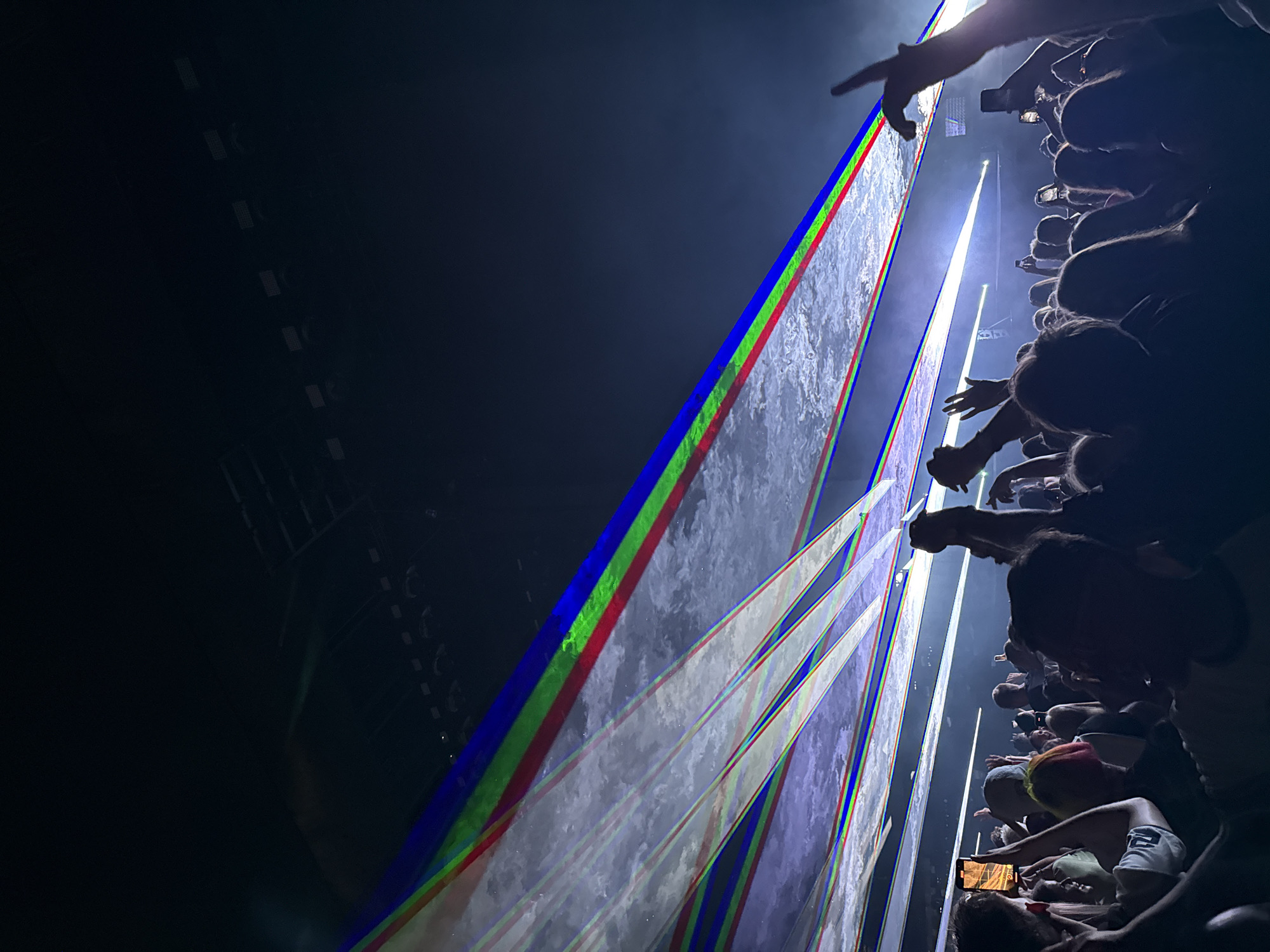 The image depicts a large crowd of people at an indoor event, likely a concert or show, with dramatic lighting effects. Laser beams, predominantly in red, green, and blue, are projected across the dark ceiling, creating a visually striking atmosphere. The crowd is silhouetted against the light, with many individuals holding up their phones, possibly to record or take pictures. Some hands are raised in the air, indicating engagement and excitement. The environment suggests a venue with a high ceiling, such as an arena or large hall. There is no discernible information in the image that places the scene in Riga, Latvia.