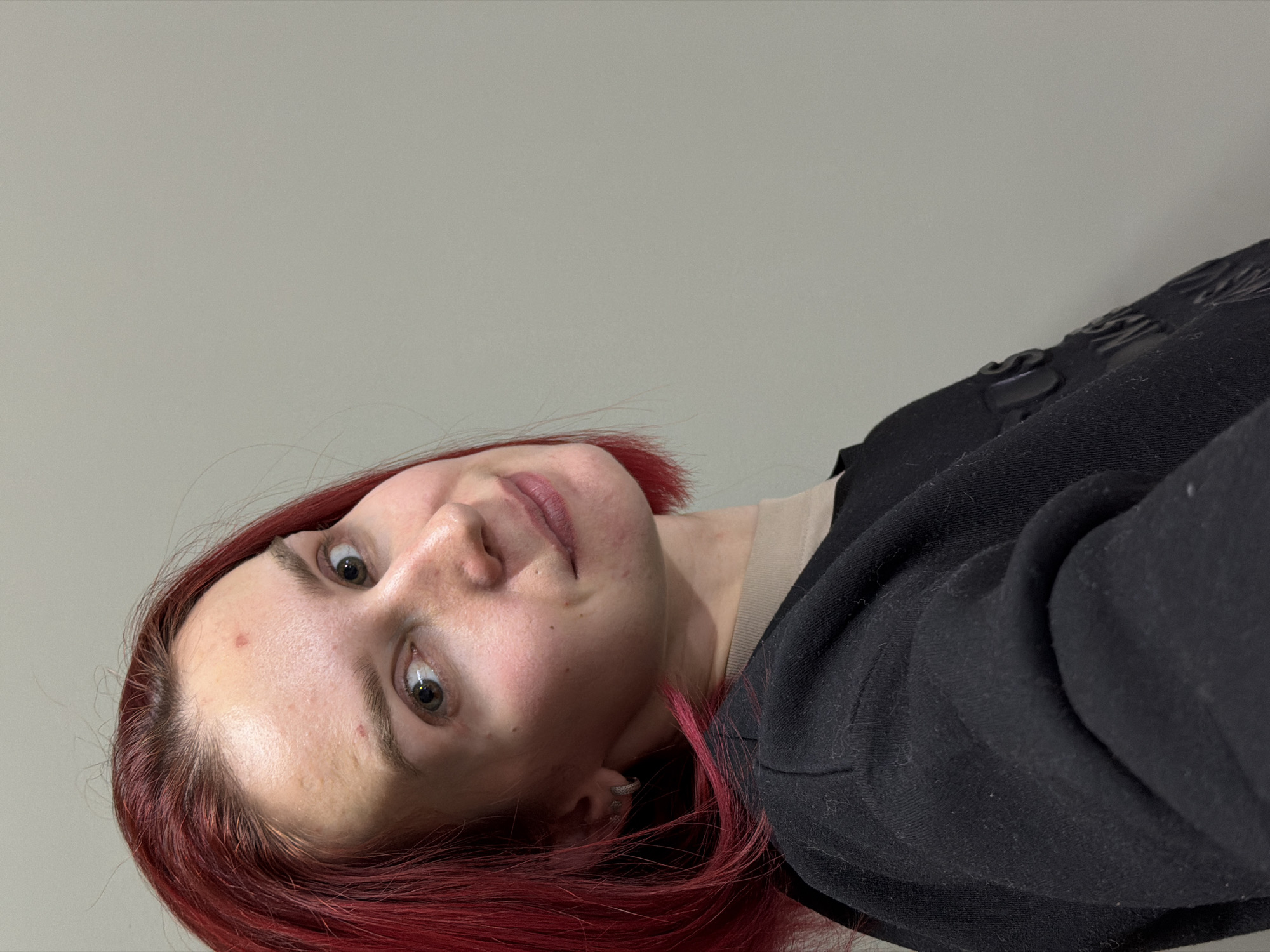 A close-up, waist-up portrait features a person with vibrant red hair, taken indoors against a plain light gray wall in Chaikovsky, Russia. The individual's head is tilted significantly to the side, and they are looking directly into the camera with wide, greenish-brown eyes. Their expression is neutral, with lips slightly parted. The skin on their forehead and cheeks shows a few small blemishes or freckles.
The person is wearing a black, casual garment, likely a jacket or hoodie, over a lighter-colored (beige or tan) undershirt. A subtle, dark embossed or stitched design, possibly an 'S' shape, is visible on the upper shoulder/arm area of the black fabric. A silver hoop earring is worn in the visible ear. The bright red hair frames the face and cascades around the shoulders. The lighting is even and soft, suggesting an indoor setting. No visible text is present in the image.