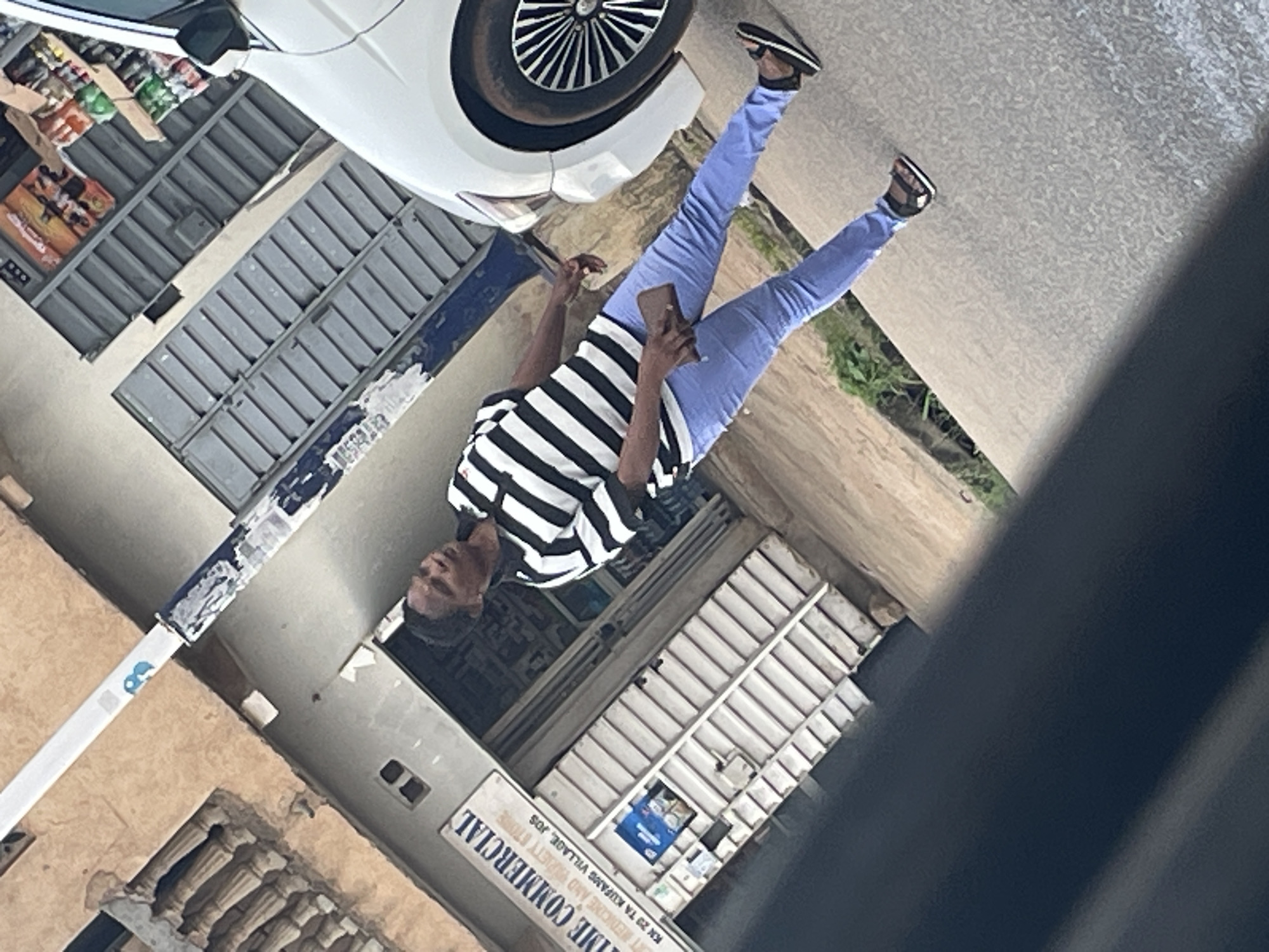 A person stands on a street in Jos, Nigeria, next to a white car and a building with a store. The person is wearing a black and white striped shirt, light blue jeans, and black sandals. The building has a sign that reads "TIME COMMERCIAL," "EXPERT MEDICINE AND VARIETY STORE," and "KN 29 TA KUFANG VILLAGE, JOS." The street is paved with concrete. The scene appears to be in a commercial area of the city.