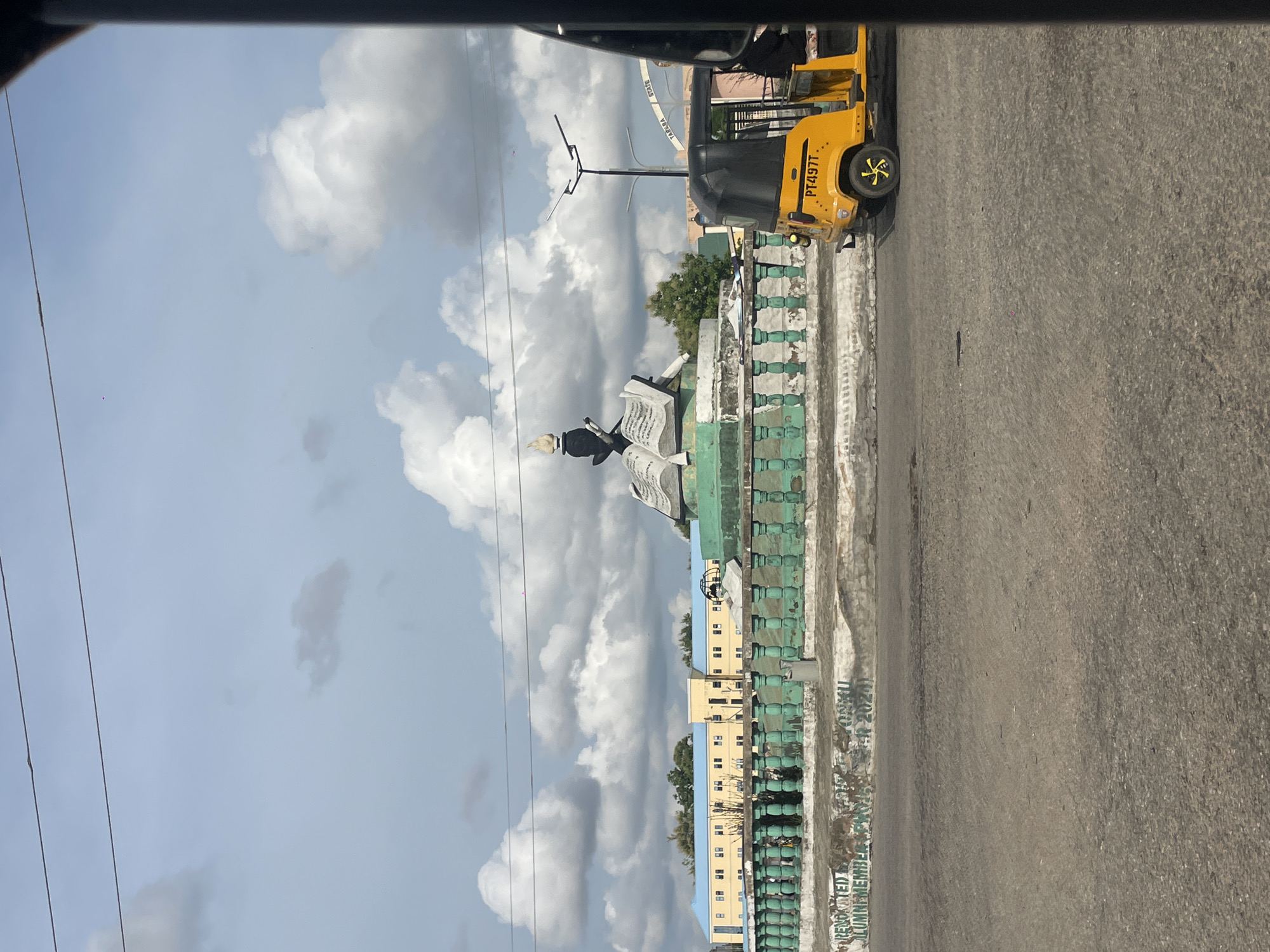 The image captures a street scene in Jalingo, Nigeria, featuring a prominent monument and a yellow auto-rickshaw. The monument, a central focus, is designed to resemble an open book with a stylized figure holding a flame on top. It is surrounded by a decorative green railing. Behind the monument, a two-story building with a blue roof and yellow facade is visible. The sky is partly cloudy, with dramatic cumulus clouds. The foreground shows a paved road, and the yellow auto-rickshaw, identified by the license plate "PT497T", is parked on the right side of the image. A person is seated in the driver's seat of the auto-rickshaw. The overall impression is of a well-trafficked urban area.