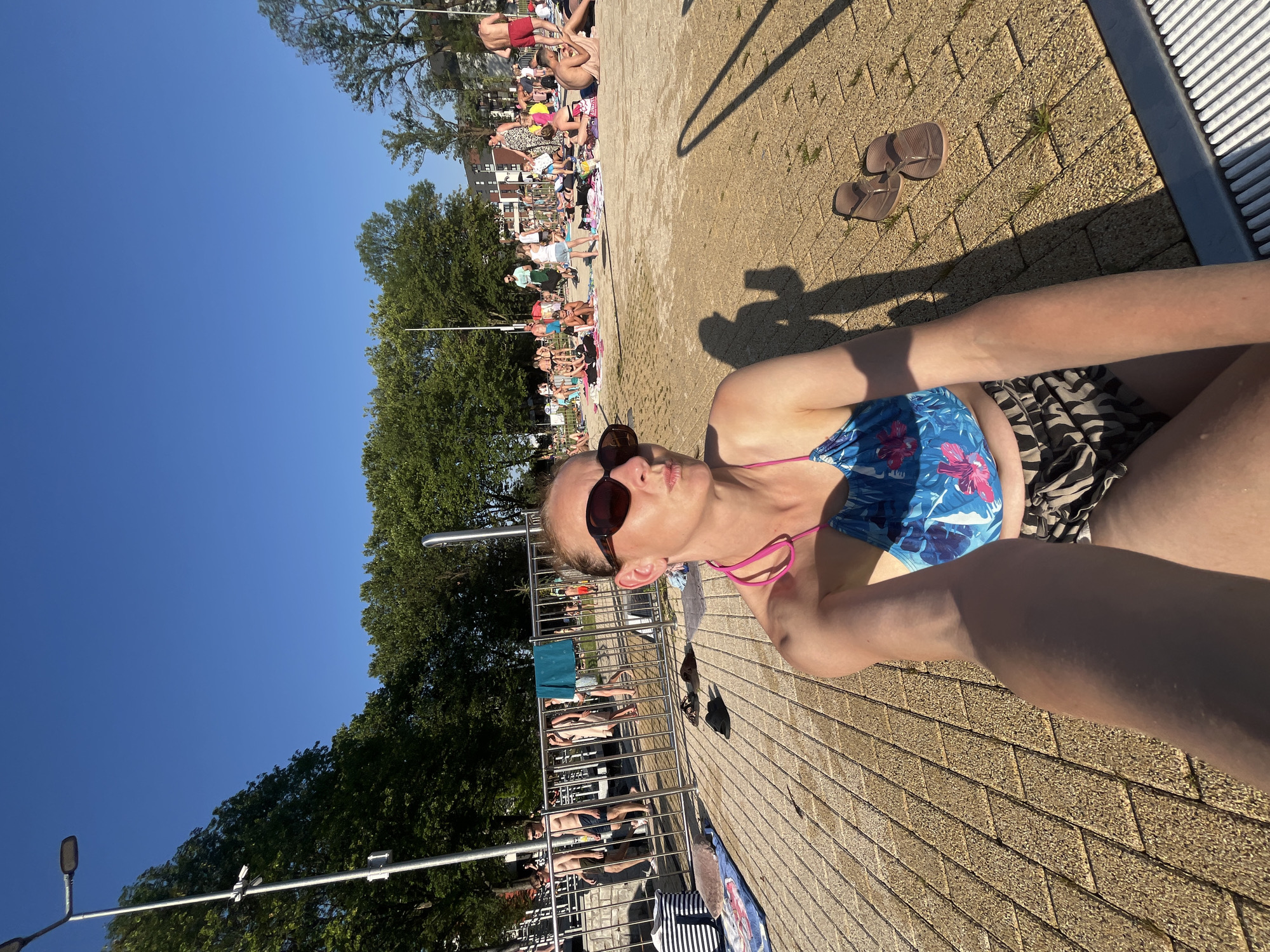 An outdoor scene captures a woman in sunglasses and a floral bikini top, sitting on a tiled ground. She is holding a camera in her left hand, taking a selfie. Behind her, a metal fence separates the tiled area from a grassy space filled with people sunbathing and swimming. The background features trees and buildings under a clear blue sky. The lighting suggests it is late afternoon.