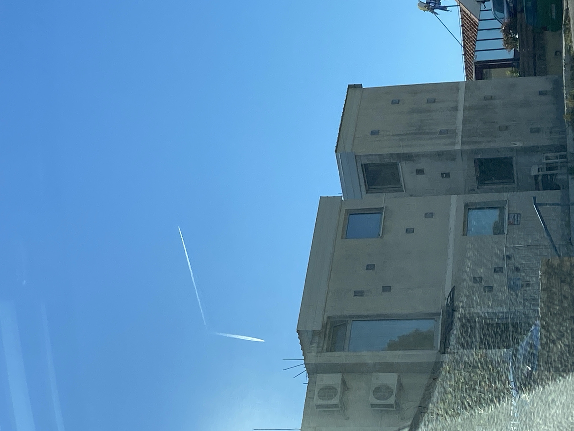 The image, taken from inside a vehicle, shows a clear blue sky over Budva, Montenegro. A contrail from a passing airplane is visible in the sky. To the right of the frame is a modern, multi-story building with a concrete facade, windows, and air conditioning units mounted on the exterior. The scene is bright and appears to be taken during daylight hours.