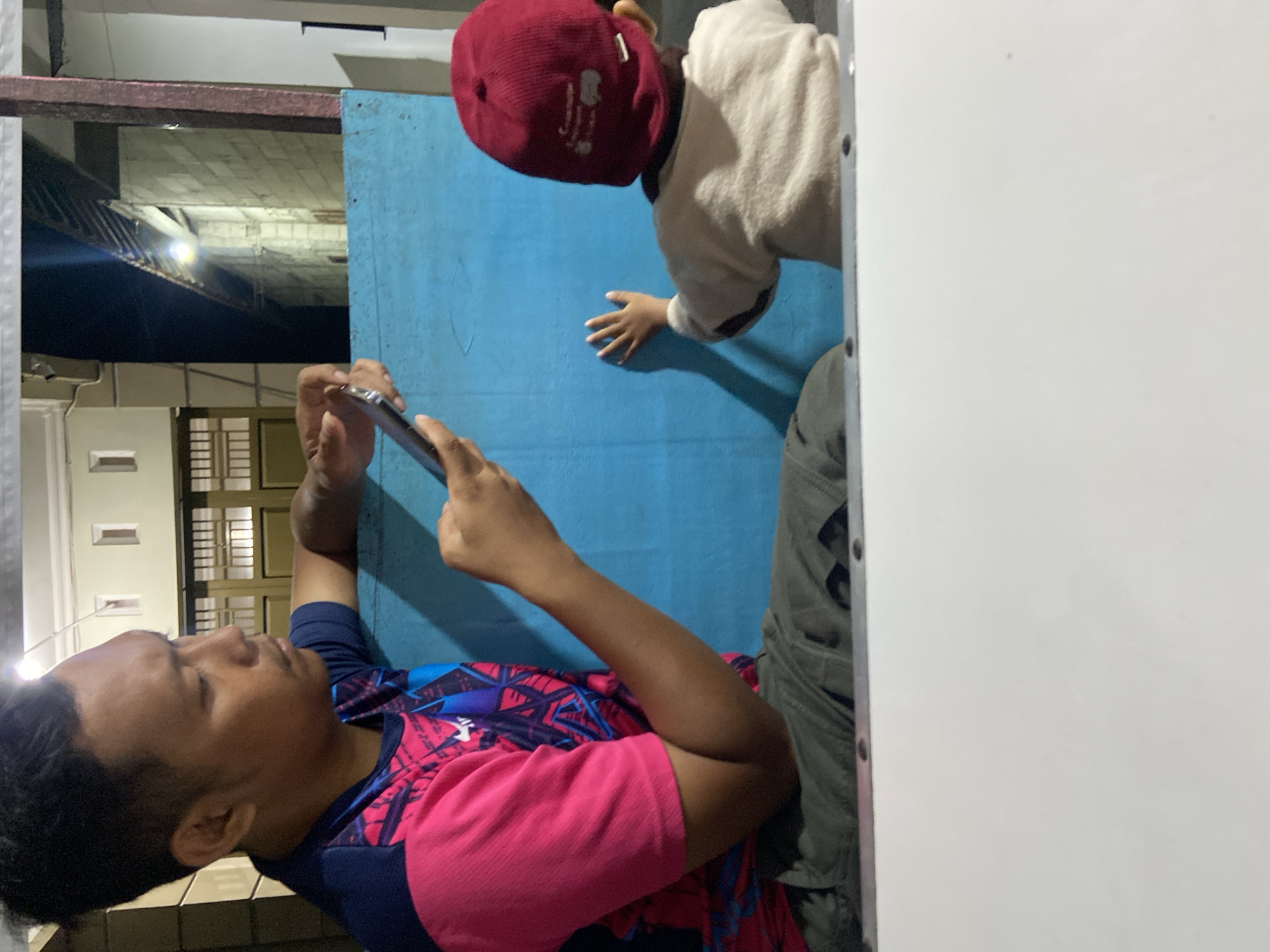 In Kecamatan Pati, Indonesia, a man in a bright pink and blue shirt is seated, holding a smartphone and interacting with a young child who is facing away from the camera. The child, wearing a cream-colored sweater and a red cap, is reaching out towards a blue surface. They are situated outdoors, with a dimly lit building facade and construction in the background, suggesting a residential or commercial area. A white surface in the foreground partially obscures the lower half of the scene.