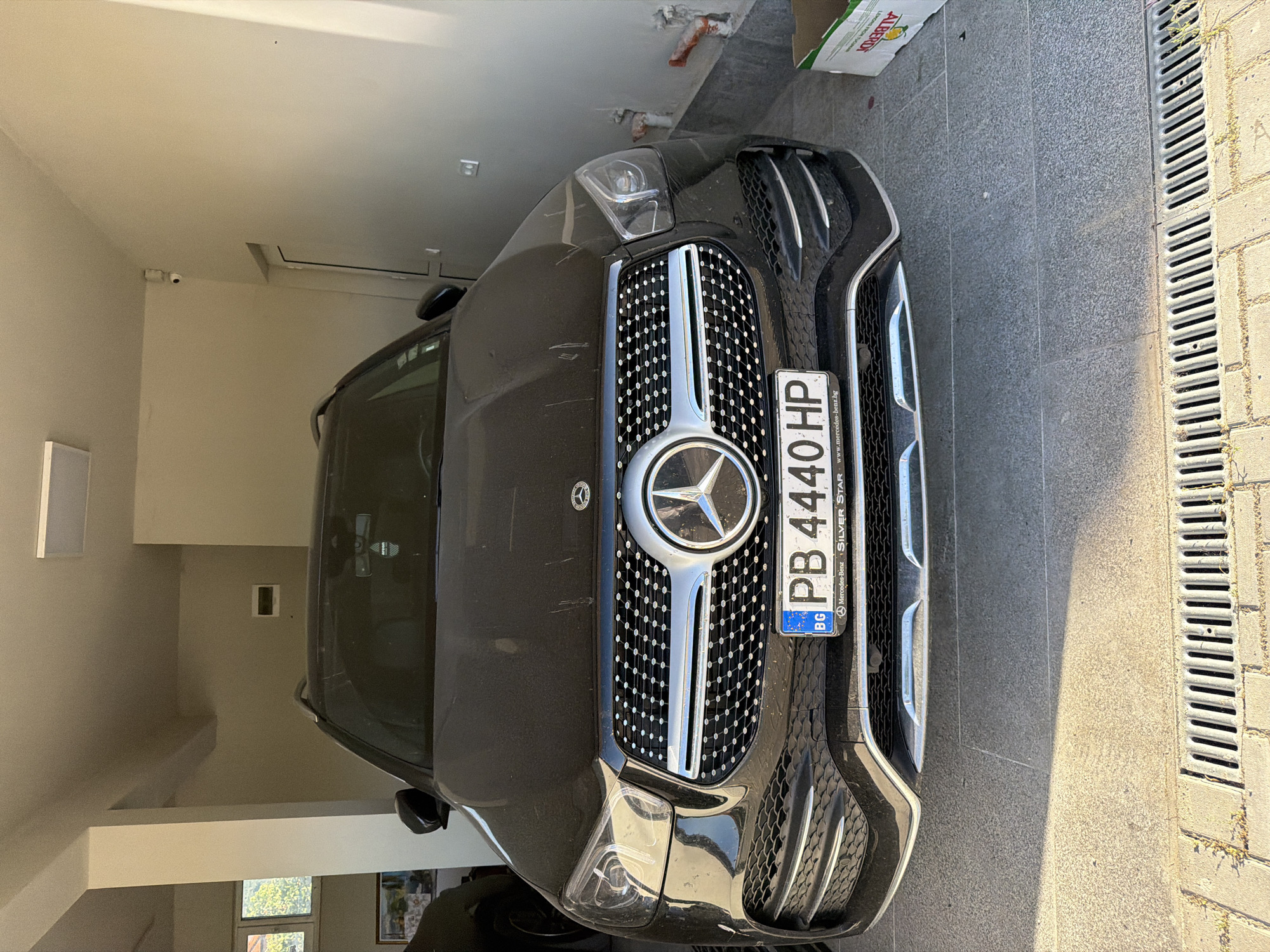 The image shows the front of a black Mercedes-Benz GLE-Class parked on a tiled surface with a drainage grate visible. The car has a Bulgarian license plate reading "PB 4440 HP." The car is parked in a garage. In the background, a surveillance camera is visible on the wall. The image was most likely taken in Asenovgrad, Bulgaria, indicated by the license plate.