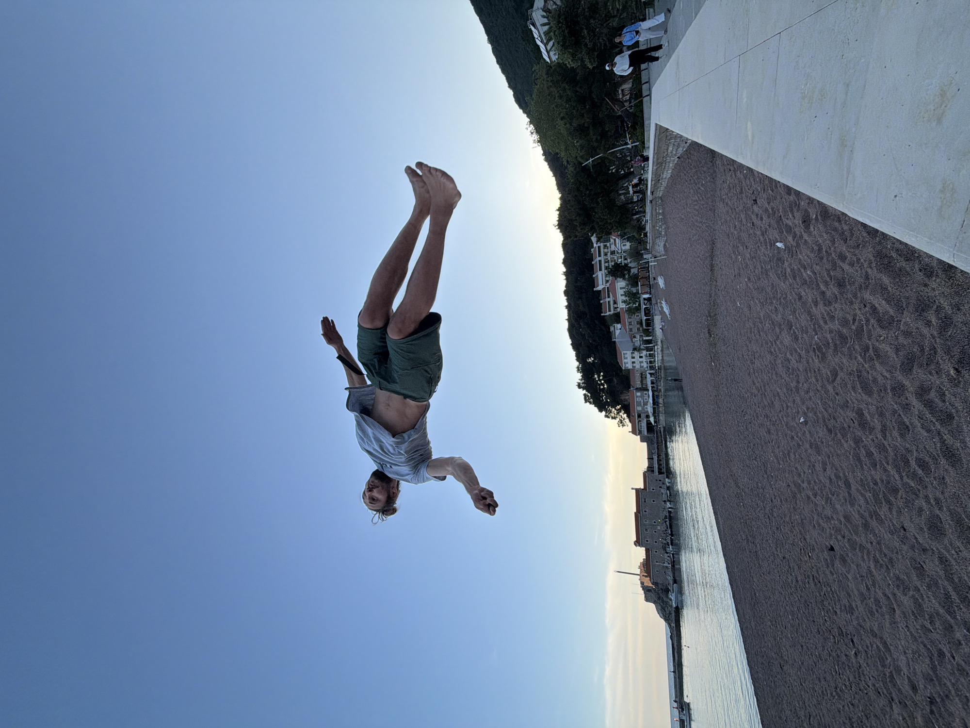 The image shows a man performing a backflip. He is in mid-air with his head pointing down and legs up. The background features a beach with dark sand, a waterfront promenade, buildings, and a mountain covered with trees. It is daytime, and the sky is clear. The location is Petrovac centar, Montenegro.