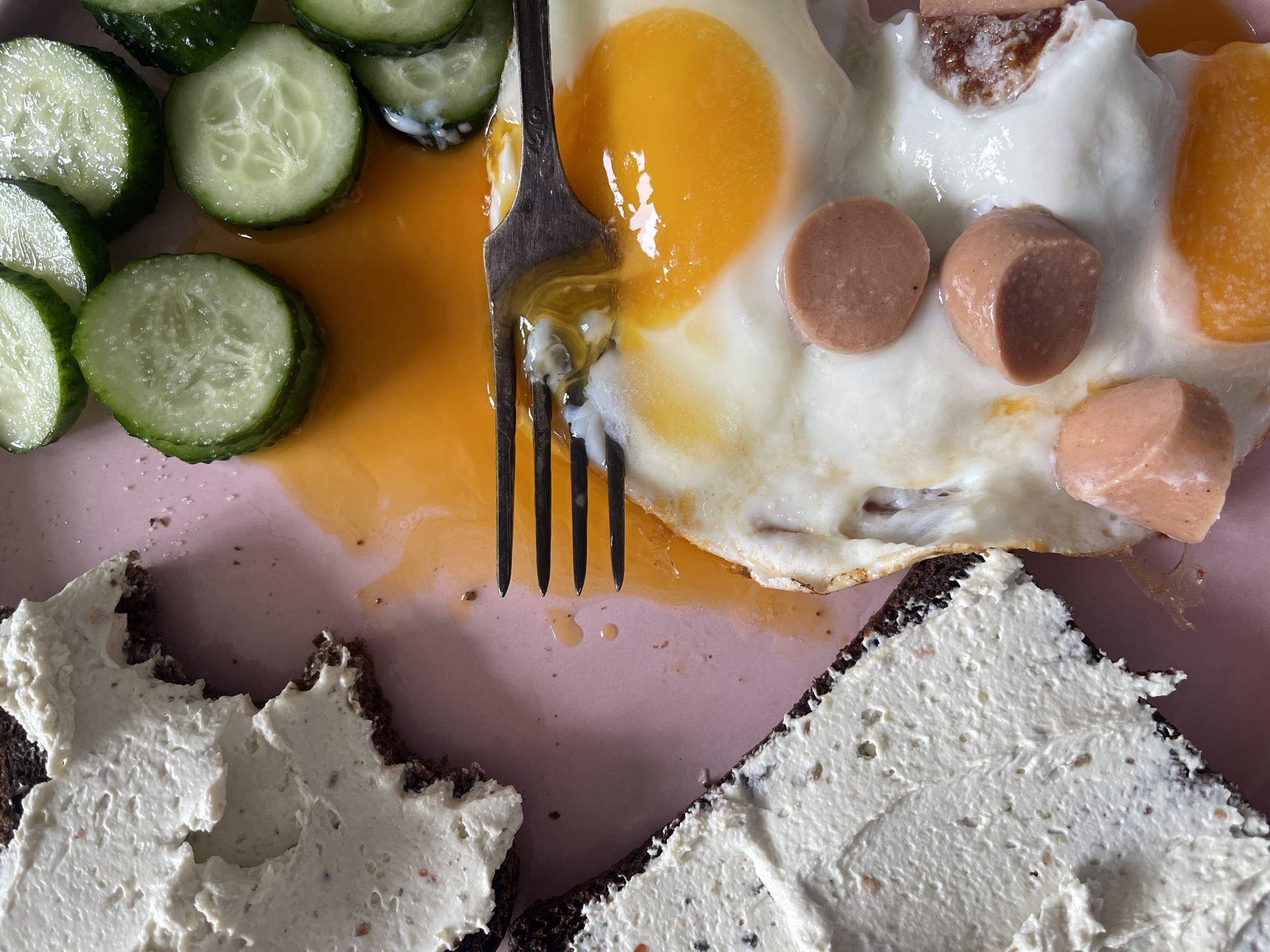 An overhead, close-up shot captures a rustic meal on a light pink plate, likely in Khimki, Russia. The main subjects are a fried egg with a bright, runny yolk that has spread across the plate, and several slices of cooked sausage. Complementing the main items are fresh, round slices of cucumber, some glistening with moisture. Two pieces of dark bread, generously topped with a thick, light-colored creamy spread, occupy the lower portion of the plate. A silver fork is prominently positioned, having just broken into the fried egg's yolk, indicating the meal is being eaten or has just begun. The lighting suggests an indoor setting, and the composition focuses on the textures and colors of the food. No people or visible text are present, but the arrangement conveys a simple, hearty breakfast or brunch.