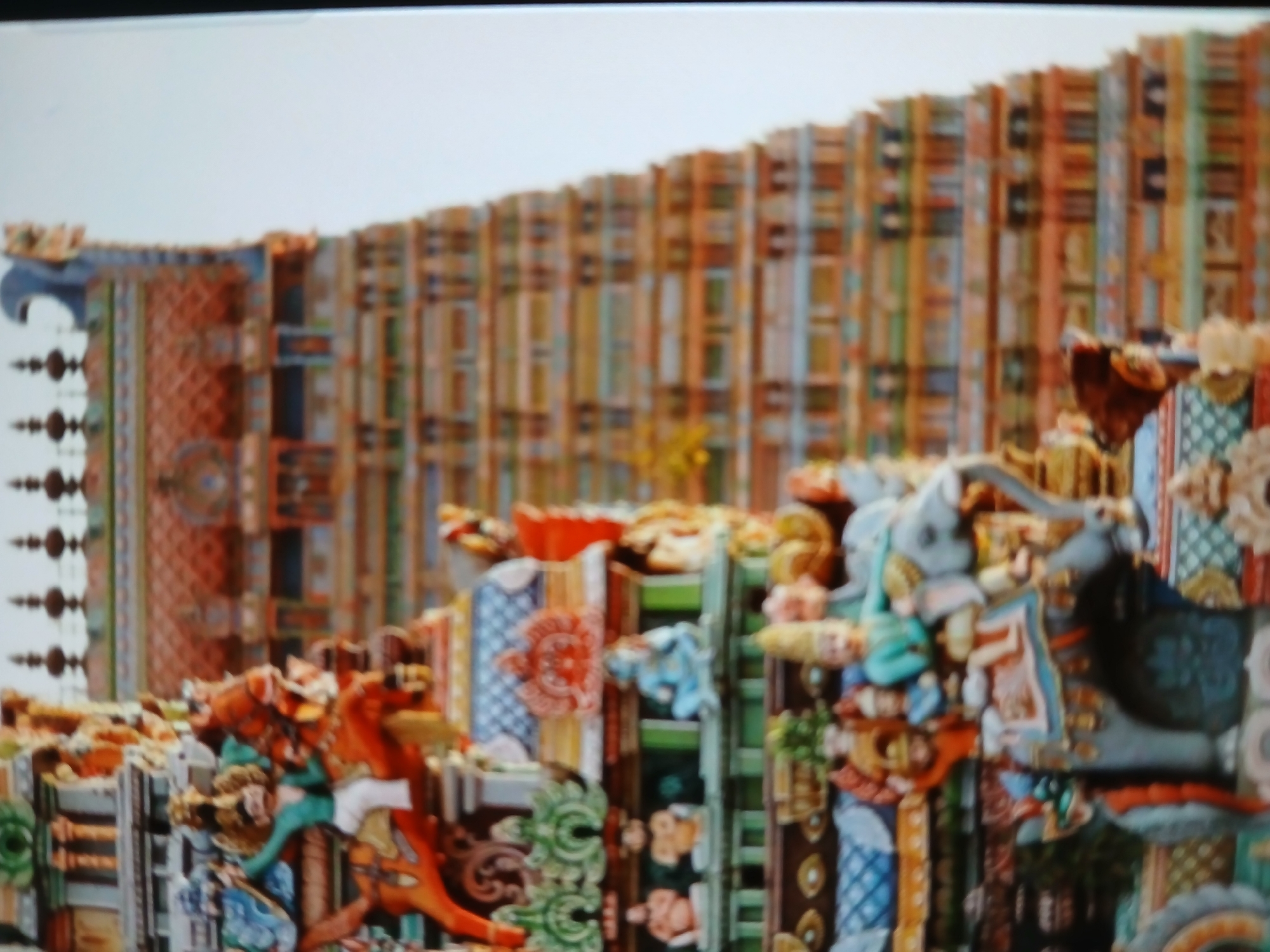 The image depicts a section of a colorful, intricately decorated Hindu temple. The structure is composed of numerous vertical segments, each adorned with painted carvings of deities, animals, and geometric patterns. The color palette is diverse, featuring shades of orange, blue, green, red, and white. The background is a dense arrangement of similar vertical elements, creating a complex and visually rich texture. The image lacks people or immediate environmental context. Specific location in Bengaluru is not discernable from the provided image.