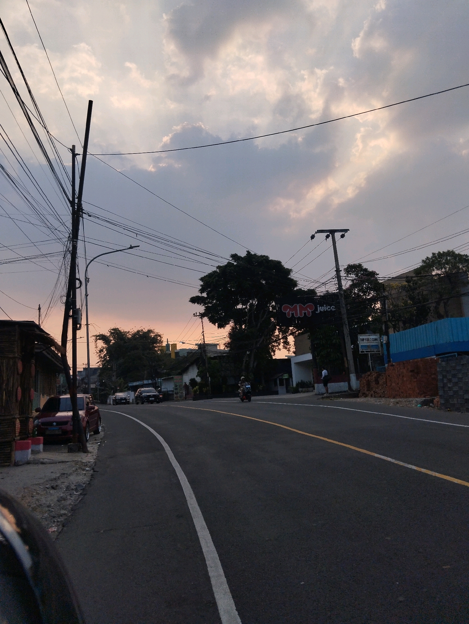 This is a vertical, medium-shot, eye-level photograph taken in Ciawi, Indonesia, during either dusk or dawn, judging by the warm, diffused light illuminating the sky and the transition to dim outdoor lighting. The scene depicts a street with a gentle curve to the right, lined with buildings and trees. On the left side of the road, there is a low-rise building with a rustic, bamboo-like exterior, and a parked red car. Further down the street, other vehicles and a motorcycle are visible.
Dominating the left side of the frame is a complex network of overhead electrical wires, supported by multiple utility poles. A prominent sign for "MM Juice" is visible across the street. The road itself is paved and features white and yellow lane markings. In the background, residential and commercial structures can be seen, interspersed with lush green trees. The sky is filled with a mix of soft gray and light orange clouds, suggesting the presence of the sun just below the horizon. A few individuals are present, including one standing near a building on the right and a person riding a motorcycle. The overall impression is that of a quiet, somewhat busy, street scene in a semi-urban environment at the edge of the day.